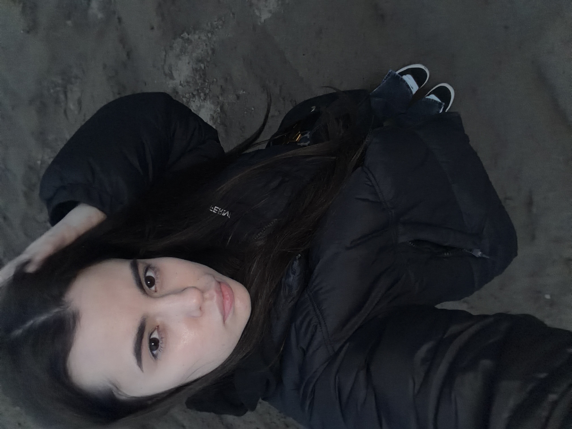 The image depicts a young woman lying or posing on a dark, textured ground surface, possibly dirt or gravel, with scattered lighter patches that may be snow or ice. She is the central subject, looking directly up at the camera. She has long, dark brown hair, which is partially fanned out around her head, with one hand gently touching it. She is dressed in a dark, likely black, puffer jacket, which suggests cold weather. A white logo or text, possibly "EEWA!" or similar, is visible on the jacket's left sleeve. She is also wearing blue jeans or trousers and black and white sneakers. The photograph is taken from a high, overhead angle. The dim lighting indicates it was captured during the evening, at night, or under very low light conditions. Her facial expression is neutral. The scene is outdoors, set in Noviy, Russia.