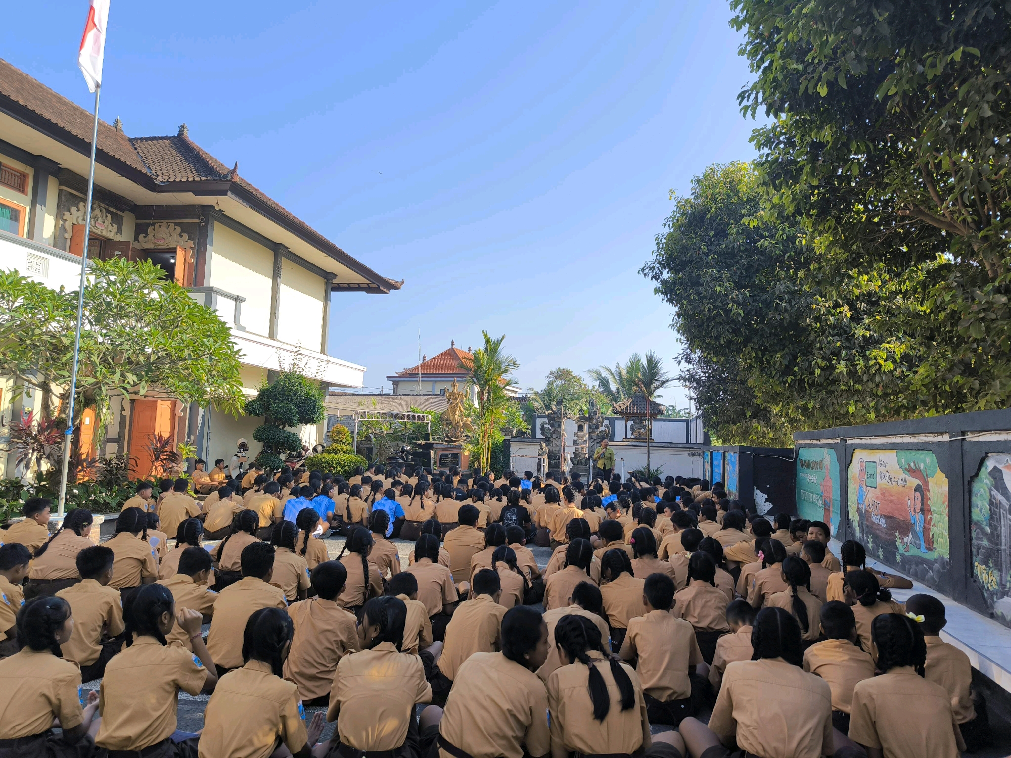 In Tabanan, Indonesia, a large group of students, predominantly wearing tan uniforms, are seated on the ground outdoors. They are facing a man in a yellow shirt who appears to be addressing them. Some students are also wearing blue shirts. The setting suggests a school assembly or event. Behind the students, a building with traditional Balinese architectural elements, including a tiled roof and decorative carvings, is visible. Tall trees and lush greenery surround the area, and a clear blue sky is overhead. The scene is illuminated by natural sunlight.