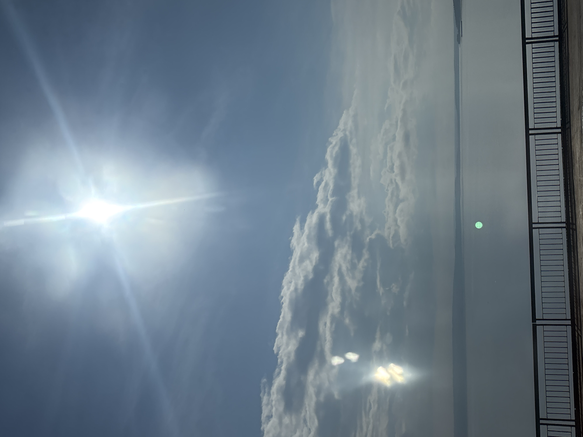 The image depicts a wide body of water under a bright, cloudy sky. The sun is visible as a strong, lens-flared light source in the upper center of the frame, casting bright rays and lens flares. Wispy white clouds are scattered across the sky, some appearing denser and more defined. In the lower portion of the image, there is a dark railing with vertical bars, suggesting a balcony, bridge, or elevated viewpoint. Beyond the railing, the water stretches to a distant horizon, where a faint landmass is visible. The overall scene is a landscape with natural elements and a man-made structure in the foreground. This image does not contain any people or specific actions. The location is not identifiable from the visual information provided in the image.