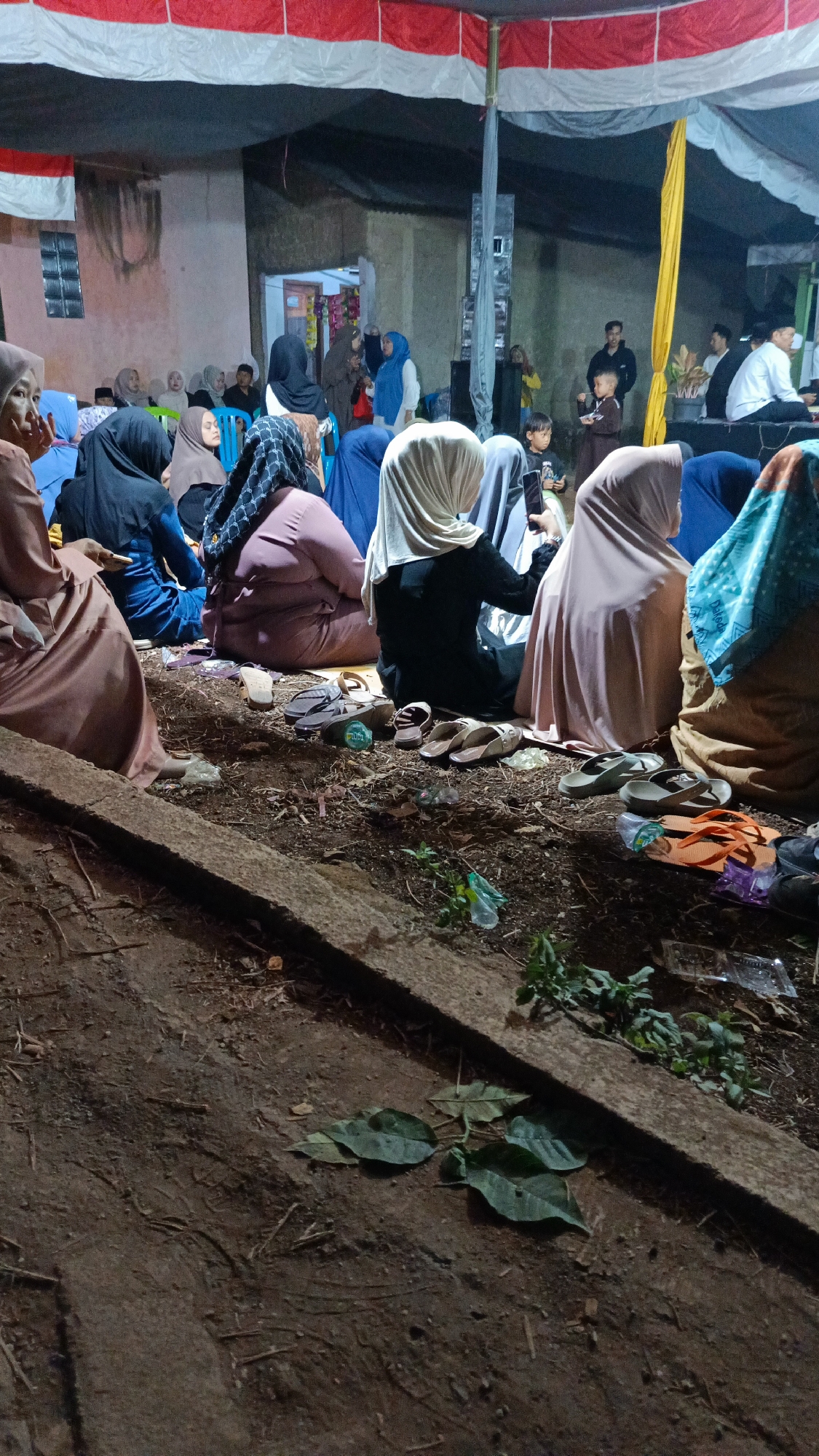 The image depicts an outdoor gathering in Margajaya, Indonesia, likely a community event or a religious service, indicated by the presence of individuals dressed in modest attire, including headscarves. The scene is set under a tent structure with red and white fabric draped overhead, suggesting a temporary arrangement. A group of people are seated on the ground, facing towards the center of the gathering. Some individuals are actively engaged, such as the person holding a mobile phone. Various footwear, including sandals and flip-flops, are scattered on the ground near the seated individuals. The environment appears to be unpaved, with dirt and debris visible. In the background, there are structures that suggest buildings or temporary stalls, and other individuals can be seen standing or seated further away, participating in the event. The lighting suggests it is evening or nighttime, with artificial light sources illuminating the area.