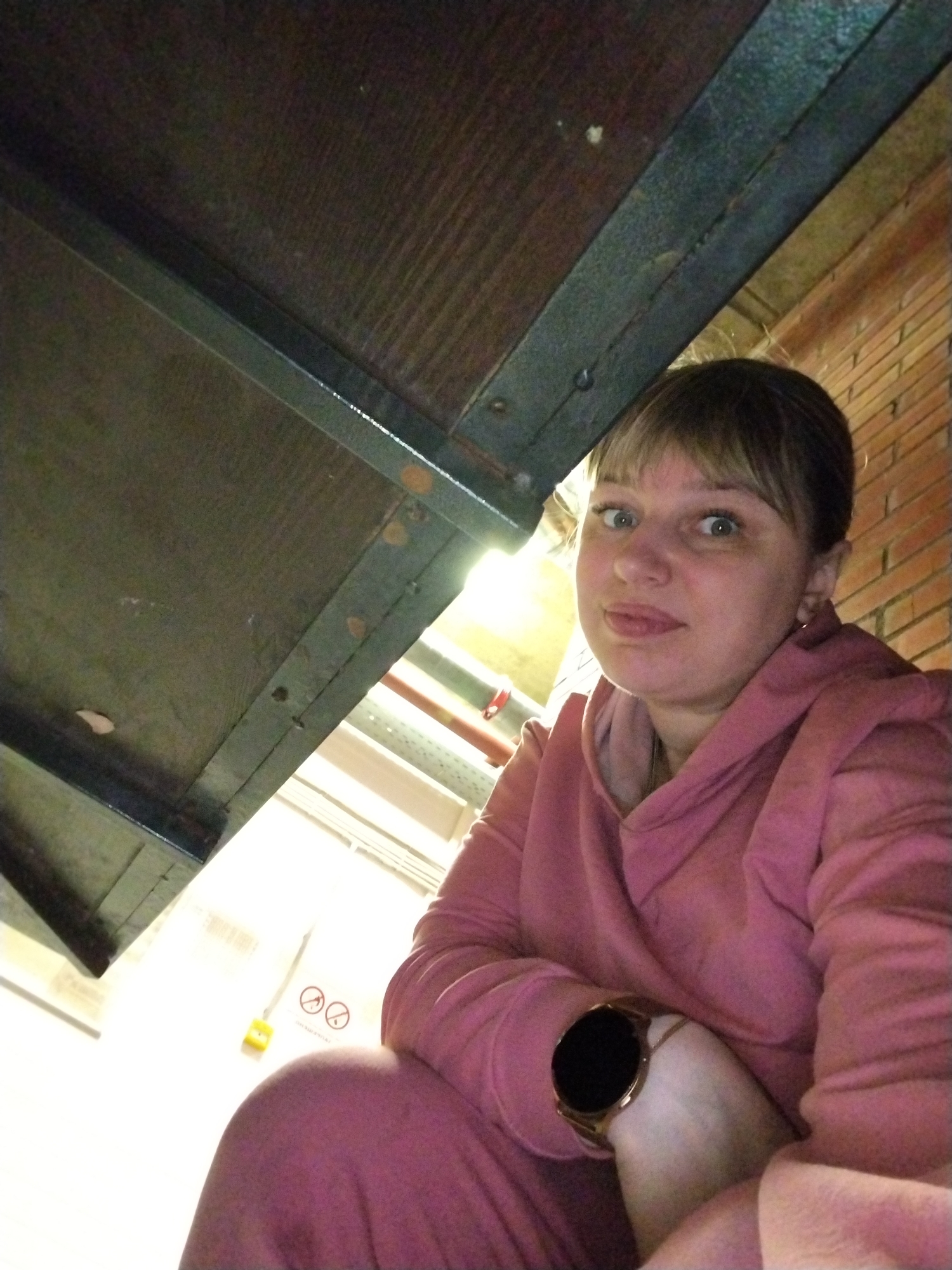 The image features a woman captured from a low-angle perspective, giving the impression that she is sitting or crouching under a large, dark, flat surface. This dominant surface, visible in the upper left, appears to be the underside of a wooden or metal structure with visible supports and screws.
The woman has light-colored hair with bangs and blue eyes, looking directly at the camera with a neutral, slightly wide-eyed expression. She is dressed in a casual pink hoodie and matching sweatpants. A smartwatch with a gold-colored bezel is prominent on her left wrist.
In the background to her right, a brick wall is visible. The area behind her left shoulder is brightly lit, revealing what appears to be artificial lighting, some red pipes, and a yellow box. Next to this box, two universally recognized "no smoking" symbols are clearly visible on a white surface. The setting suggests an indoor environment, possibly a basement or an older industrial-style building. The activity is a direct-gaze portrait or selfie. The location context is Шебунино, Russia.