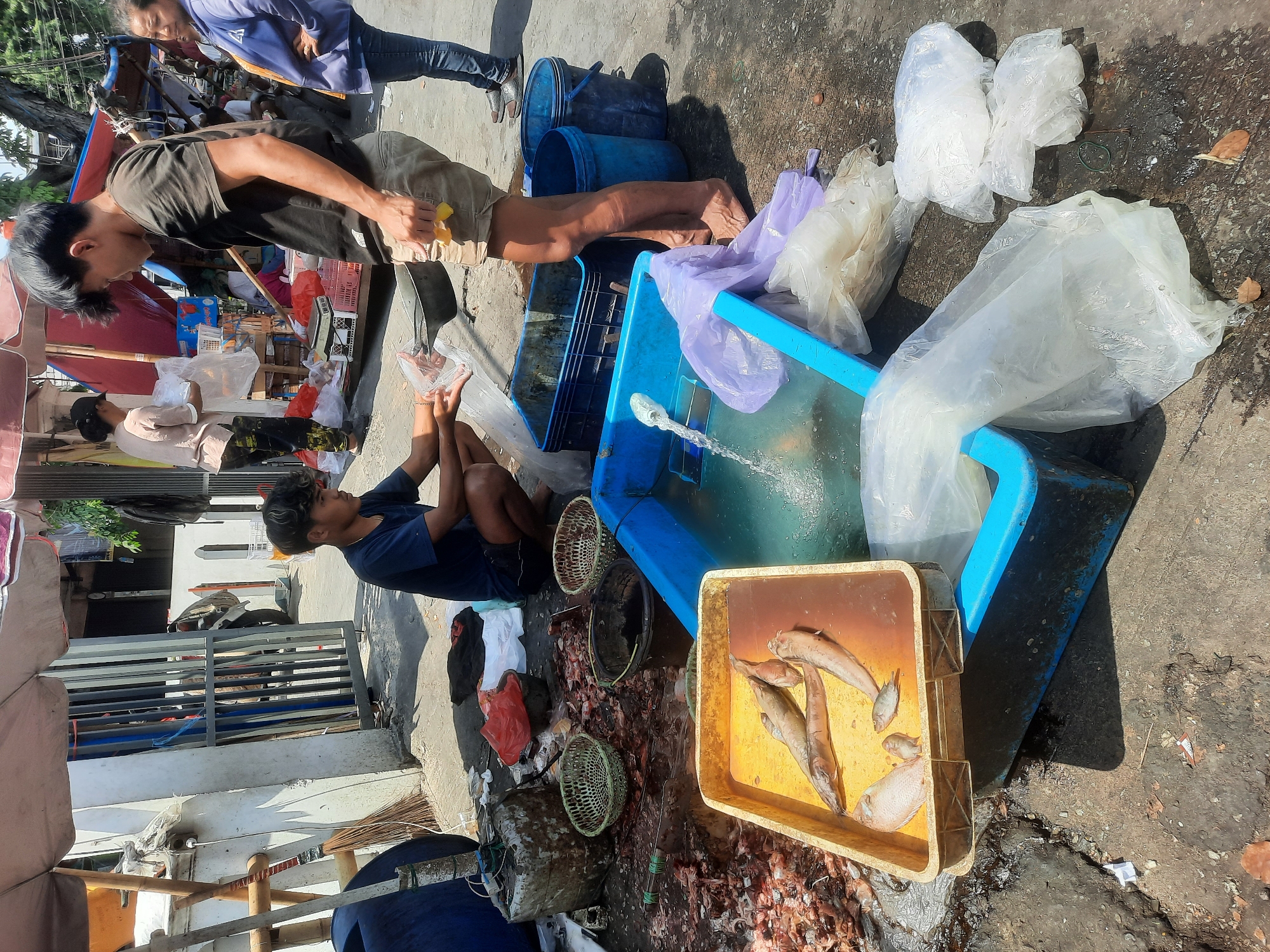 The image depicts an outdoor scene, likely a street market, in North Jakarta, Indonesia. Two young men are the central figures. One, wearing a dark t-shirt and khaki shorts, is standing and appears to be engaged in a transaction, holding a yellow object. The other, wearing a dark blue t-shirt and dark shorts, is squatting and holding a clear plastic bag containing what appears to be fish. In front of the squatting man is a yellow plastic tray filled with several fish and a smaller fish. To the right of this tray is a large blue plastic container with water, a running water source, and a purple plastic bag. Several other blue buckets and plastic bags are scattered on the ground, along with discarded fish parts and remnants. In the background, other individuals and market stalls are visible under awnings. The ground is paved and appears wet in some areas.