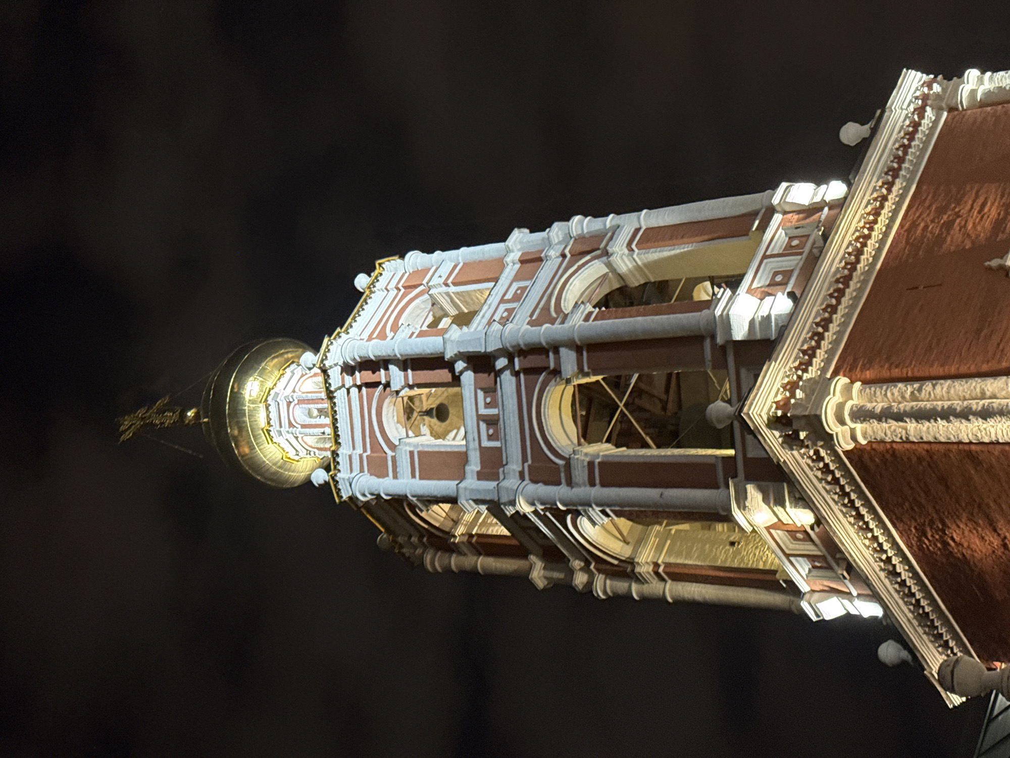 This is a low-angle night shot of a red brick bell tower with white architectural accents and a golden dome topped with a cross. The tower is illuminated by spotlights, casting light on its ornate details. The sky is dark and cloudy, typical for a nighttime scene in Moscow, Russia. The architectural style appears to be Orthodox Christian, common in the city. No people or other objects are visible in the frame.