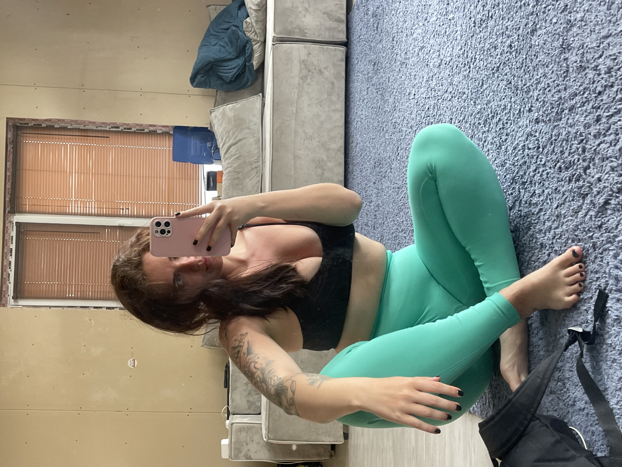 A woman is taking a selfie in what appears to be an indoor setting. She is seated on a blue carpeted floor wearing a black sports bra and green leggings. Her dark hair is down, and she has a tattoo on her left arm. A black bag is visible near her feet. Behind her is a grey couch, a window with blinds, and a wall that appears unfinished. It is not possible to determine the location in the city of Мосметаллоконструкция Онт, Russia from the image.