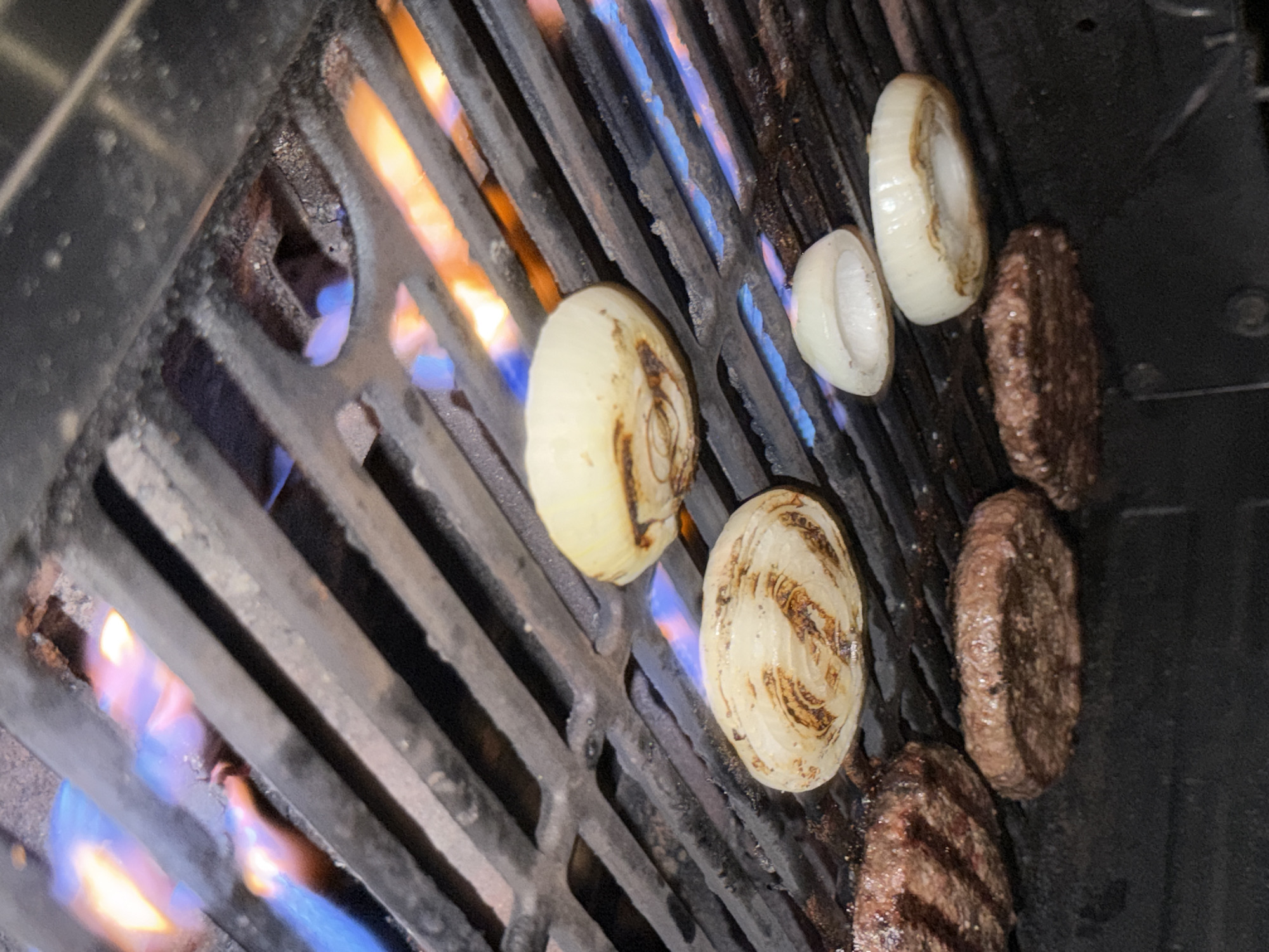 This image captures a close-up view of a grill with several hamburger patties and rings of onion being cooked over open flames. The grill grates are dark and appear well-used, with visible char marks and remnants of food. Flames, predominantly blue and orange, are visible beneath the grates, indicating active cooking. There are four hamburger patties visible, all appearing to be browned and cooked on one side, with some showing grill marks. Three rings of white onion are also present on the grill, with one ring appearing to be slightly charred and caramelized. The scene suggests an outdoor cooking event, possibly in a residential backyard or a park, given the nature of the equipment. The lighting suggests it might be evening or nighttime, enhancing the visibility of the flames. The location is identified as Hampton Bays, United States of America.