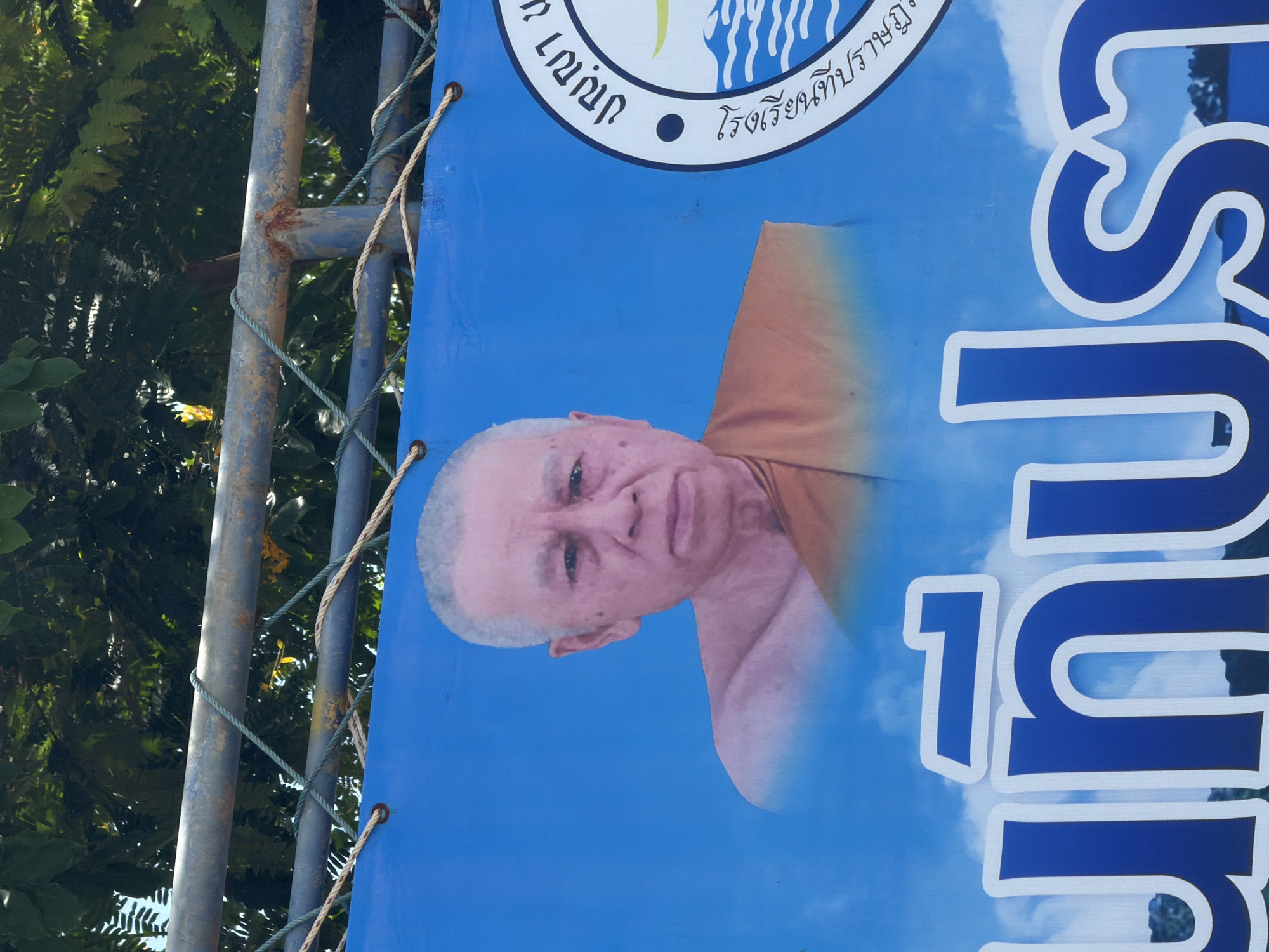 The image is a close-up shot of a blue banner displaying a portrait of an elderly man in saffron robes. The man has a shaved head with white hair, wrinkled skin, and appears to be looking directly at the camera. The banner is strung up outdoors, with a rusty metal railing and green foliage visible in the background. There is also text in Thai script on the banner, and a circular emblem with more text and an image of waves. The banner seems to be advertising an event or a religious figure associated with Baan Bo Phut, Thailand. The overall impression is a public display, likely in a temple or a community area.