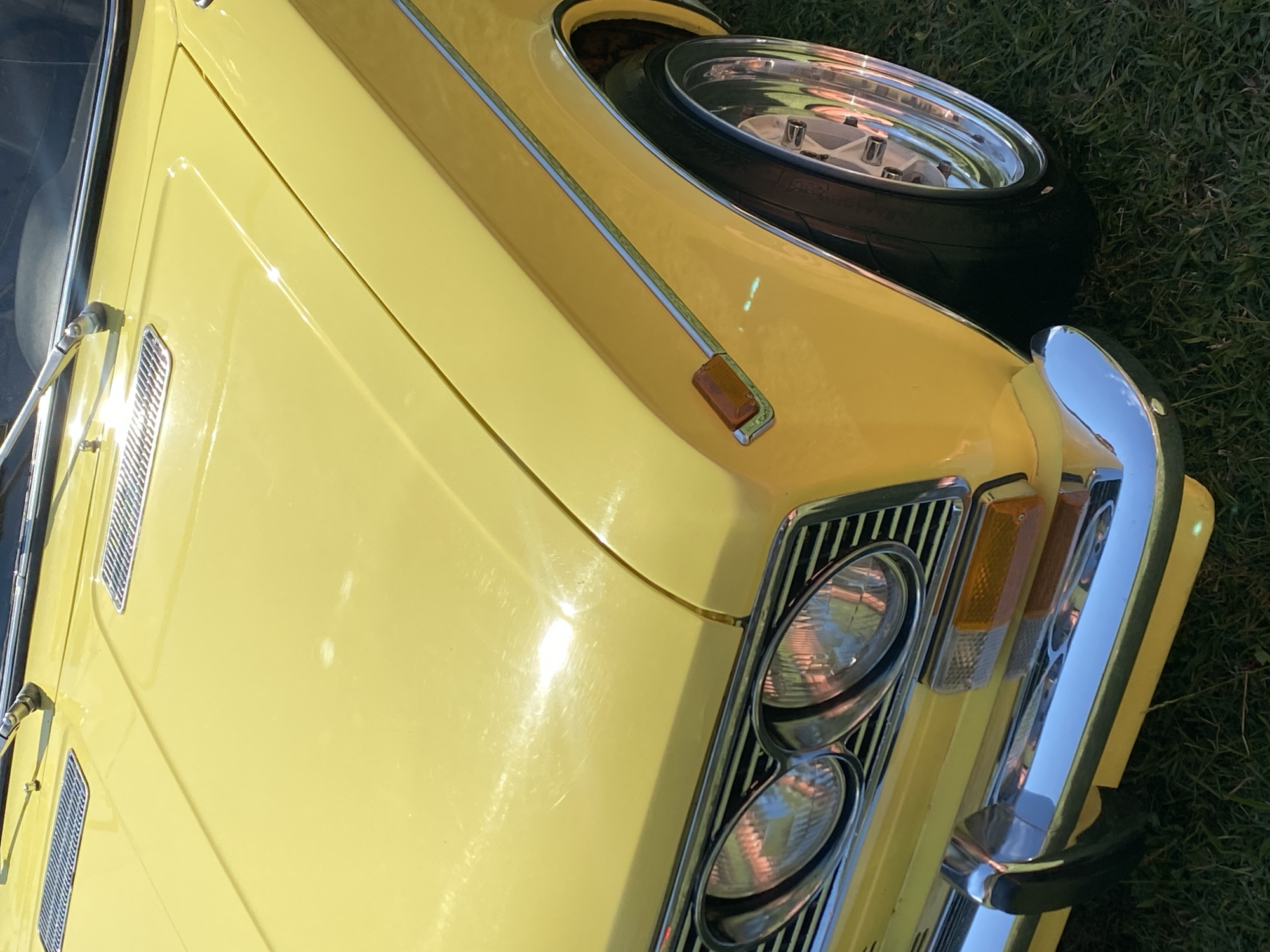 A close-up, eye-level shot captures the front quarter panel and hood of a bright yellow classic car parked on a grassy area, possibly in Bezymyannoe, Russia. The car's polished chrome bumper and grille gleam under natural light, reflecting the surrounding environment. Two circular headlights are visible, along with an amber turn signal on the fender. A chrome trim line runs along the car's body, meeting the wheel arch where a shiny chrome rim and black tire are partially visible. The hood features two rectangular metallic vents, and a windshield wiper is extended. The background consists of green grass, suggesting an outdoor setting. No people are present in the frame.