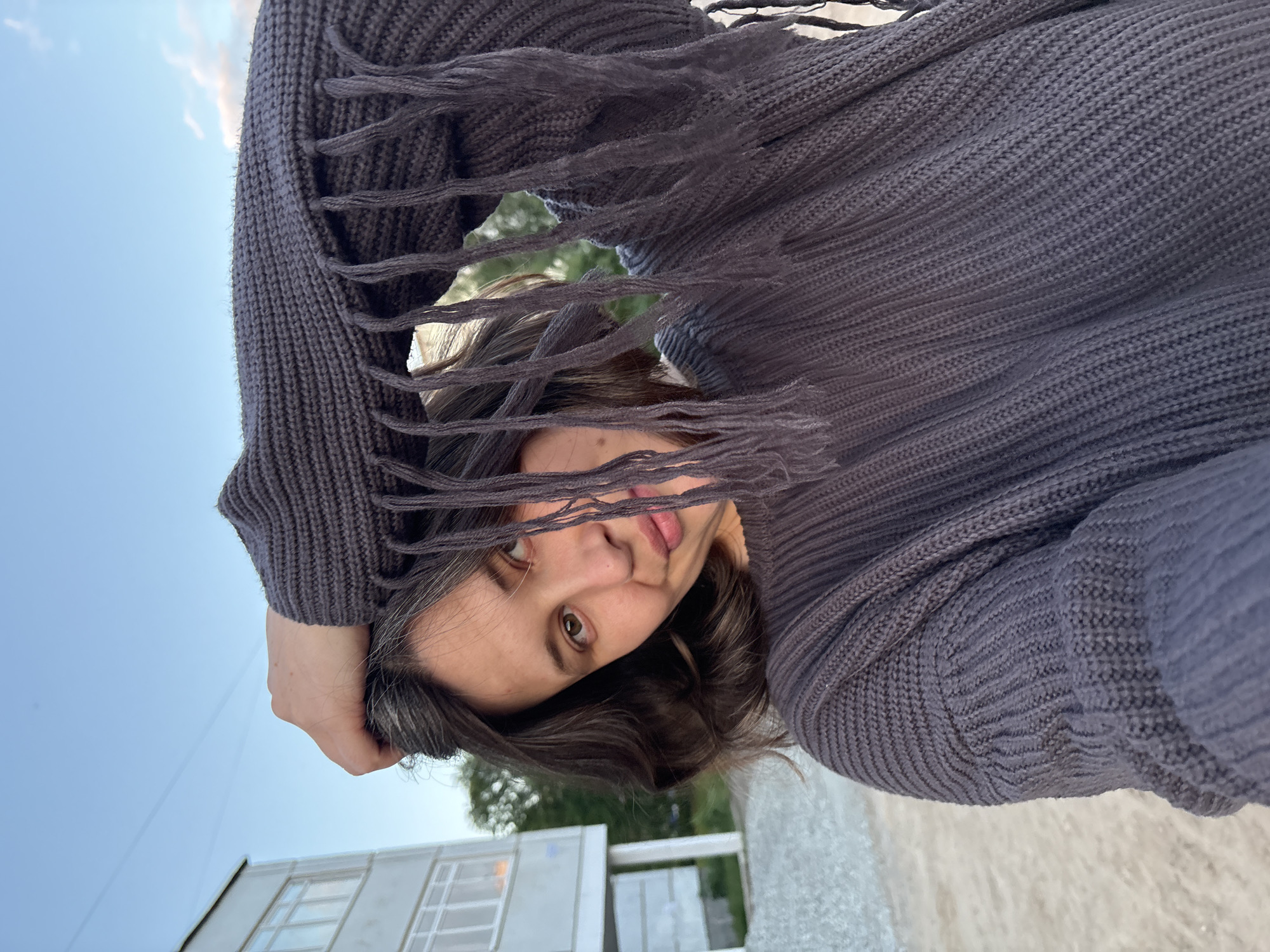 A young adult female, appearing to be in her late teens or early twenties, looks directly at the camera with a subtle smirk and brown eyes. Her dark brown, shoulder-length hair exhibits visible grey strands near the scalp on the left side of her head. She is wearing an oversized, dark charcoal grey chunky knit sweater or poncho, featuring a distinct ribbed texture on the body and sleeves, with a prominent fringe detail across the upper portion that partially obscures her face on the right side. Her left hand is raised, with fingers lightly touching her head near the crown, partially covered by the garment.
The scene is an outdoor setting in Chaikovsky, Russia, during what appears to be late afternoon or early evening. In the background to the left, a multi-story residential building with light-colored walls and white-framed windows is visible. Sparse green trees and foliage are present in the mid-ground behind the subject. The immediate foreground is a light brown, unpaved area of dirt or sand, while a darker, possibly paved or gravelly surface is visible further back to the right. The sky is predominantly clear and light blue, with faint airplane contrails visible as thin white lines in the upper left quadrant. Towards the upper right, the sky shows a softer blue transitioning to hints of pink and white clouds, characteristic of twilight.