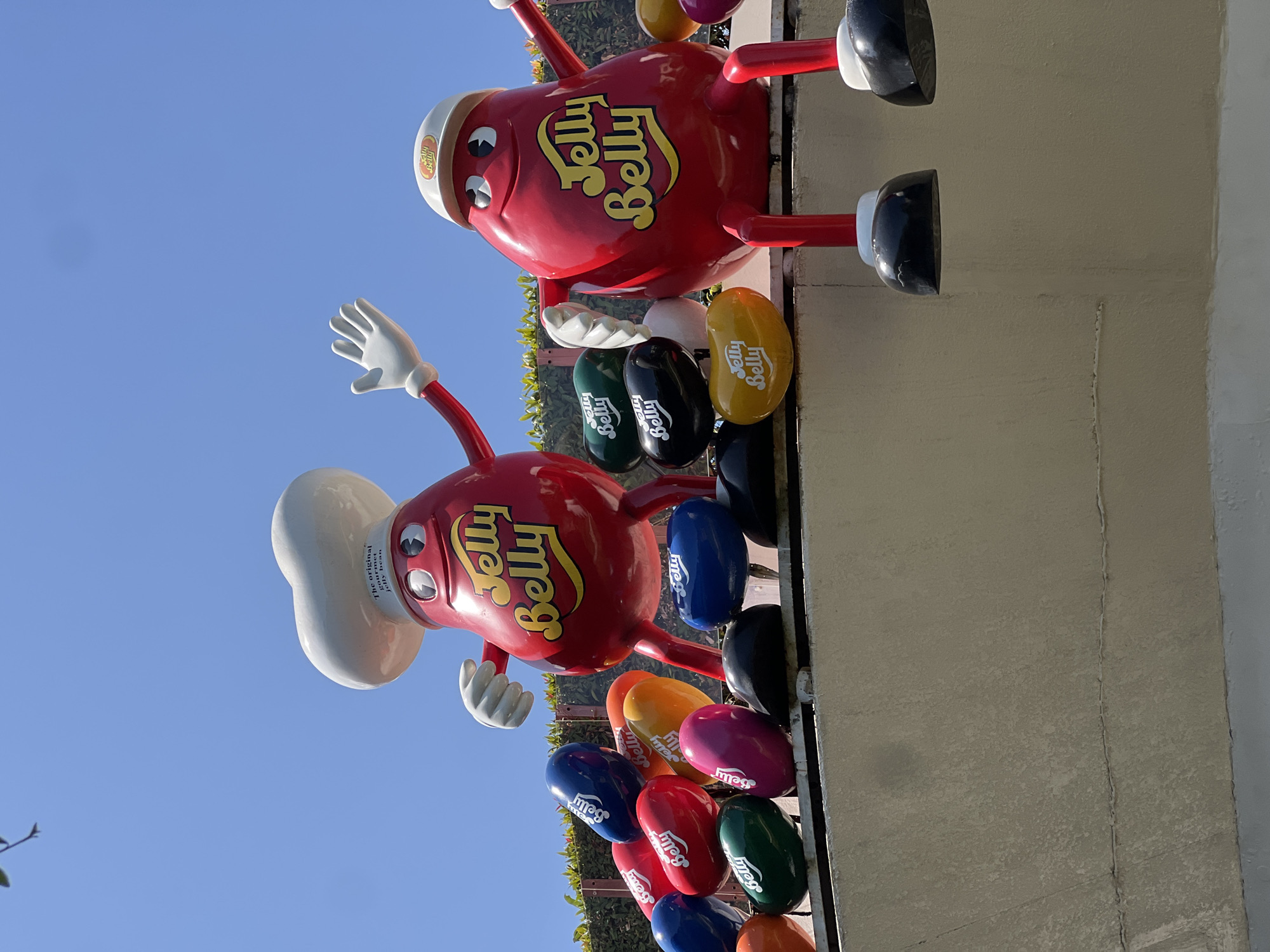 Two large, red, anthropomorphic jelly bean figures are displayed outdoors against a clear blue sky. The figure on the left wears a white chef's hat with "The original gourmet jelly bean" printed on it. Both figures have white gloves and red limbs, with black shoes. They are adorned with the "Jelly Belly" logo. Numerous large, colorful jelly bean-shaped objects with the "Jelly Belly" logo are arranged around them. The figures and jelly beans are perched on a light-colored ledge or wall. The background features green foliage and a bright blue sky. Based on available information, this display is located at the Jelly Belly Candy Company in Kuta, Indonesia.