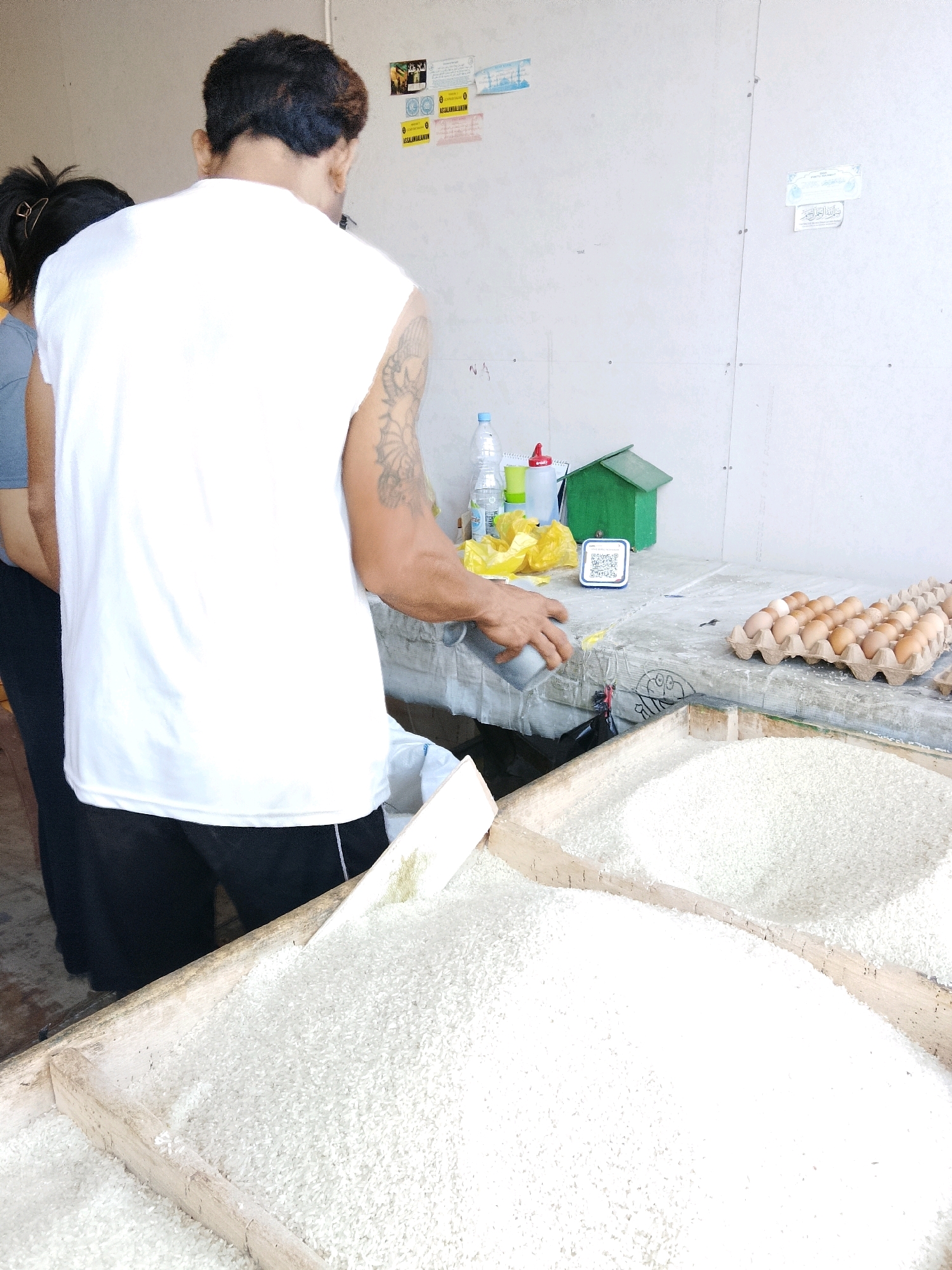 In Talise, Indonesia, a man wearing a white sleeveless shirt and black shorts stands behind a table. He is holding a gray container and appears to be scooping something into a white bag or container. In front of the table are two large wooden crates filled with white rice. To the right of the table, a tray of eggs is visible, along with a small green structure that resembles a house. A bottle of water and other items are also present on the table. The background consists of a plain wall with some papers attached to it. Another person, partially visible, stands to the left of the man. The overall scene depicts a market or shop setting where goods are being sold.