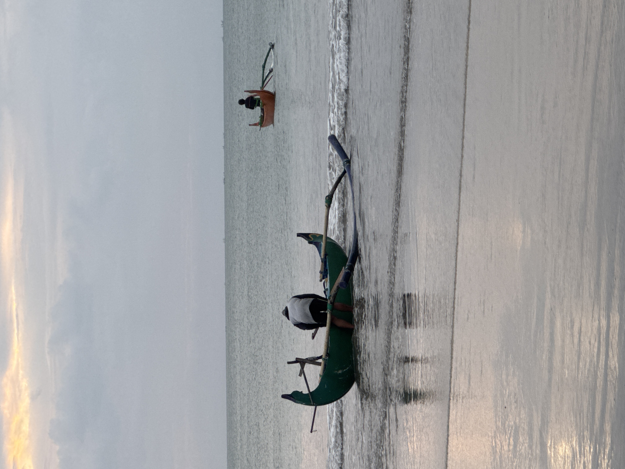 The image depicts a serene beach scene at dusk, likely in Kuta Selatan, Indonesia. The setting is a calm ocean with gentle waves lapping at the shore. The sky is overcast with a hint of warm sunset colors peeking through on the left side of the frame.
Two traditional outrigger canoes are visible. In the foreground, a green canoe rests partially on the wet sand. A person wearing a white shirt and dark shorts is crouched inside, seemingly preparing or tending to the boat. Oars are visible, one extending from the canoe. The canoe itself has a distinctive curved hull.
Further out in the water, a smaller, orange canoe is present. A person is seated in this canoe, also with oars. The overall mood is peaceful and tranquil, suggesting the end of a day of fishing or activity on the water. The wet sand reflects the muted light of the sky, creating a smooth, shimmering surface. There is no visible text in the image.