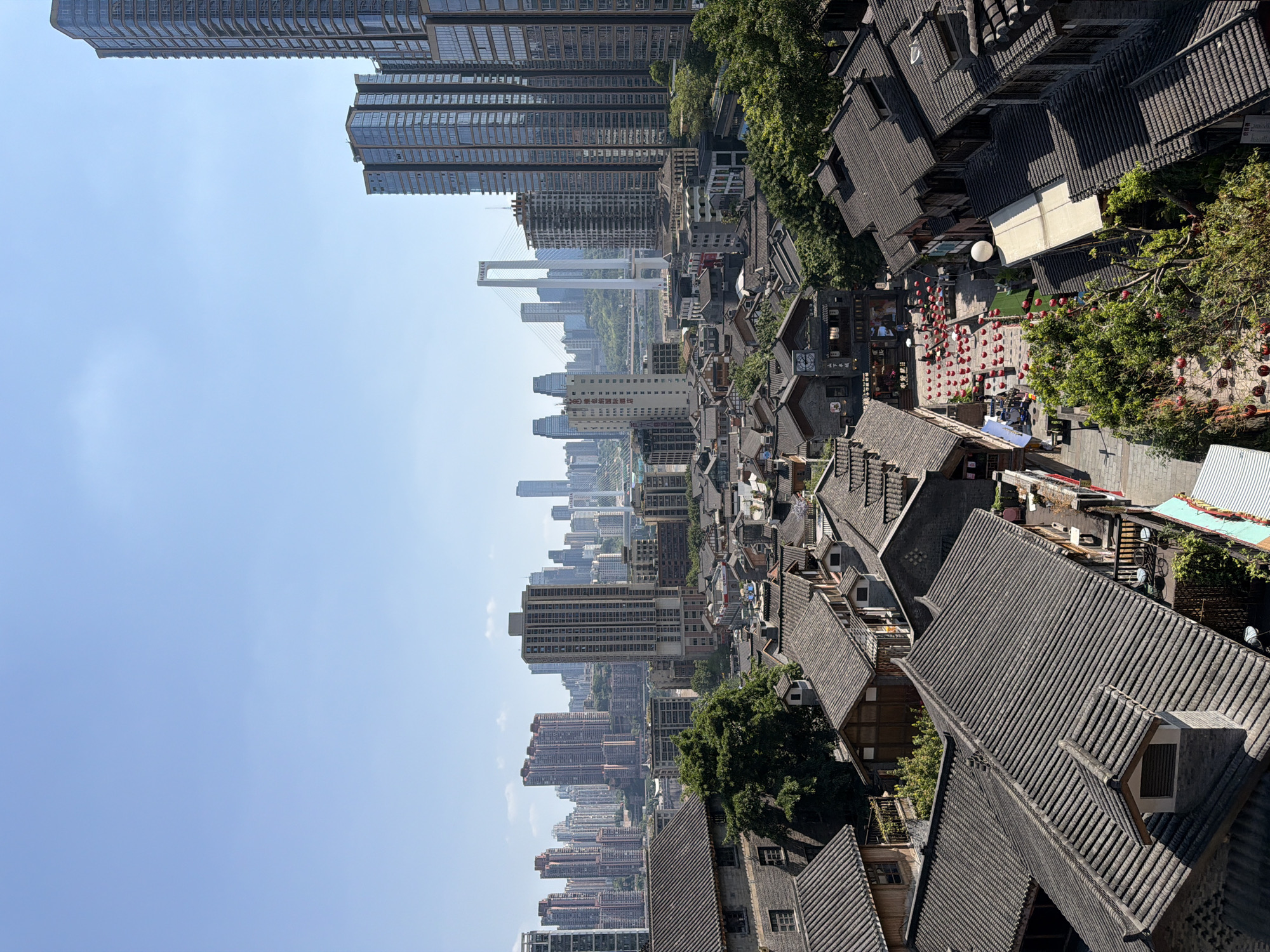 This is an elevated, horizontal, eye-level shot of a bustling Yuzhong District, China. The foreground features a dense cluster of traditional Chinese buildings with dark, tiled roofs, closely packed together. The buildings have wooden accents and are interspersed with lush green trees. A narrow street or pathway winds through this older section of the city, decorated with numerous red lanterns. Several people can be seen walking along the path.
In the midground, the traditional architecture gives way to a more modern cityscape. A collection of tall, glass-fronted skyscrapers rises prominently, contrasting sharply with the older buildings. These modern structures vary in height and design, creating a distinct urban skyline.
In the background, a large, white suspension bridge stretches across a body of water, connecting parts of the city. More skyscrapers and buildings are visible in the distance, forming a vast urban panorama under a clear blue sky with a few scattered white clouds. The overall scene depicts a blend of historical and contemporary Chinese urban development.