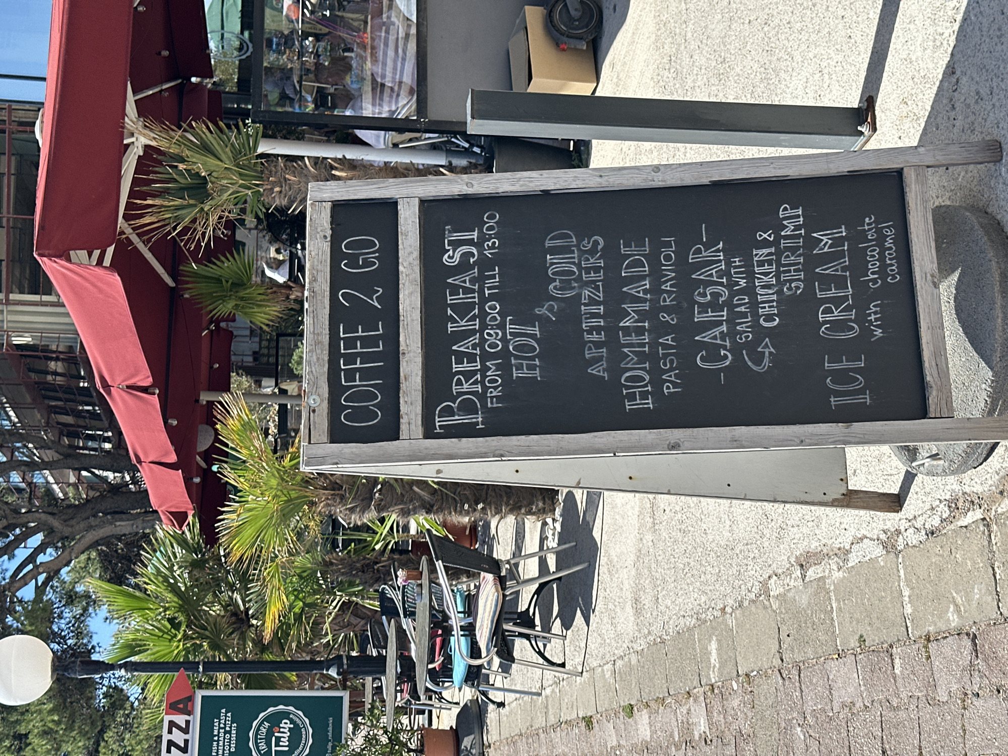 The image shows a menu board advertising "Coffee 2 Go" and breakfast available from 09:00 to 13:00. The menu includes hot and cold appetizers, homemade pasta & ravioli, Caesar salad with chicken & shrimp, and ice cream with chocolate caramel. The menu board is located outside a restaurant in Rafailovići, Montenegro. In the background, there are tables and chairs, palm trees, and a red awning. A sign for a restaurant called "Trattoria Tulip" is visible below the menu board.