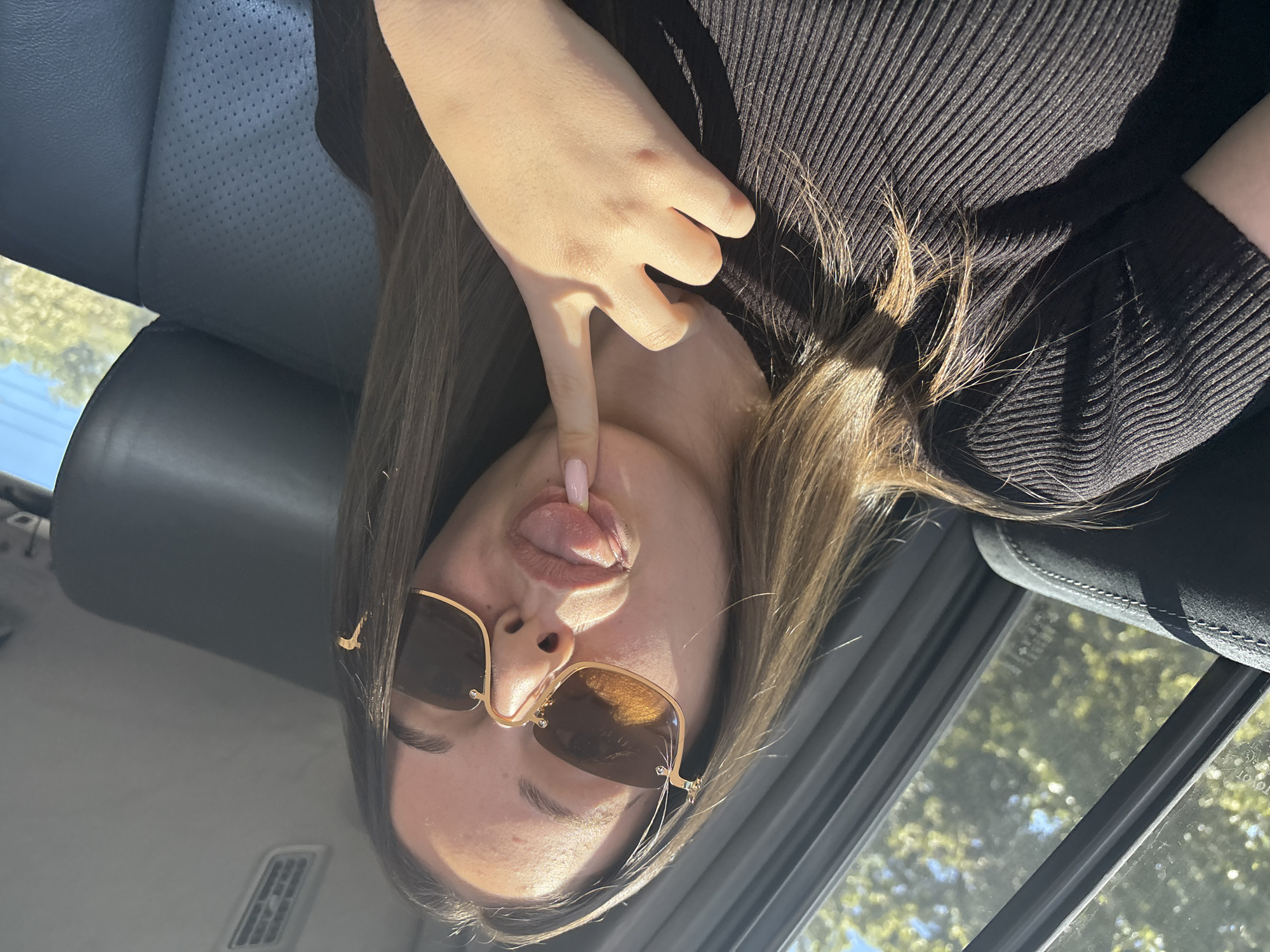 A young woman is in the front seat of a car. She is wearing sunglasses and sticking her tongue out while touching her tongue with her finger. She is wearing a black sweater and the interior of the car is gray. The location is impossible to determine from the image.