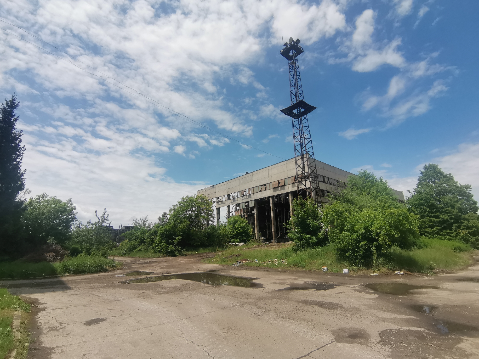 The image depicts an abandoned industrial building located in Zhivkovo, Bulgaria. The building is a large, concrete structure with many broken windows. A tall metal tower is attached to the building. The surrounding area is overgrown with trees and vegetation. The foreground features a cracked concrete surface with several puddles. The sky is blue with white clouds. There are no people visible in the image.