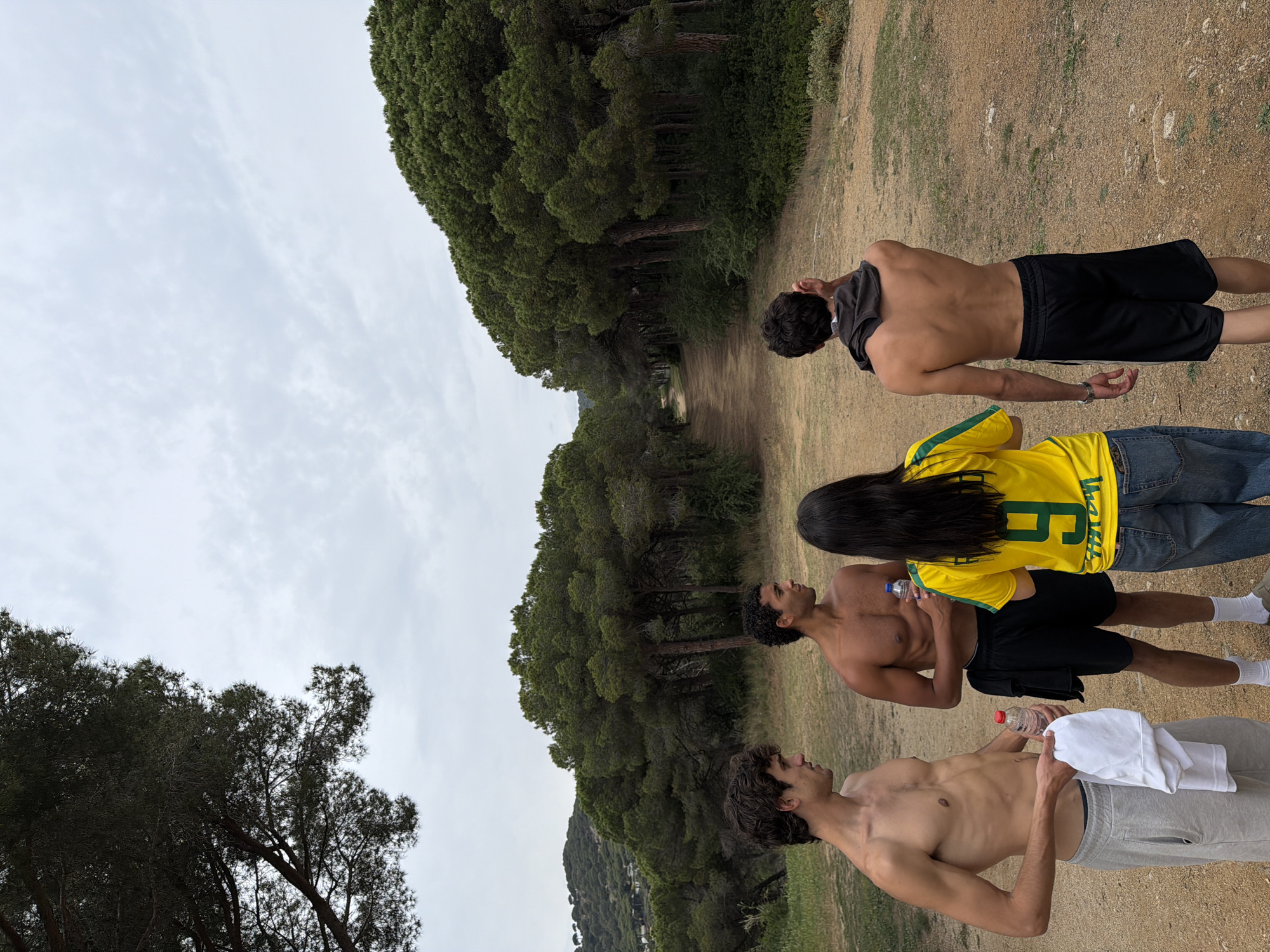 The image depicts a group of four people on a dirt path, likely in a wooded area in Sant Andreu de Llavaneres, Spain, based on the landscape characteristics. The sky is overcast.
From top to bottom, the first person is a male, shirtless, pulling a black t-shirt over his head. He is wearing black shorts.
The second person is wearing a yellow sports jersey with the number "9" on the back and blue jeans. Their long, black hair is visible.
The third person is a shirtless male holding a water bottle. He is wearing black shorts and white socks.
The fourth person is a shirtless male holding a water bottle and a white towel. He is wearing grey shorts.
All four people are standing in a row, facing the same direction, along the dirt path, which is bordered by trees. The ground is mostly dirt and grass.