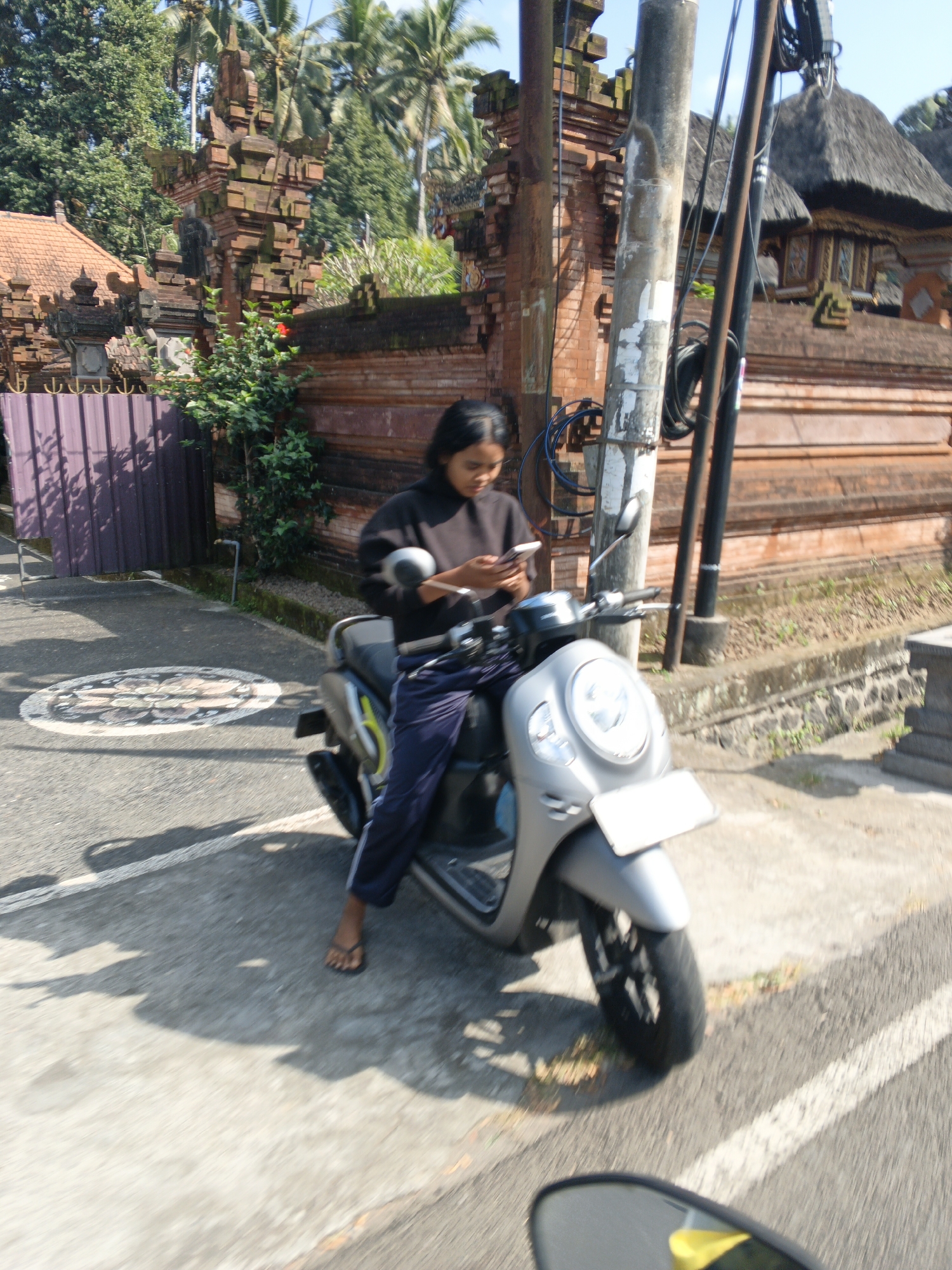 The image shows a woman sitting on a silver scooter in front of a traditional Balinese building with intricate brickwork and thatched roofs, located in Abiansemal, Indonesia. The woman is wearing a dark long-sleeved shirt and navy blue pants. She is looking down and appears to be using a smartphone. The scooter is silver and has a white license plate. The background features lush green trees, including palm trees, and a purple metal fence. The ground is paved with asphalt, and there are shadows from the scooter and overhead wires. The scene is captured in daylight with a slight motion blur, suggesting the photo was taken from a moving vehicle.