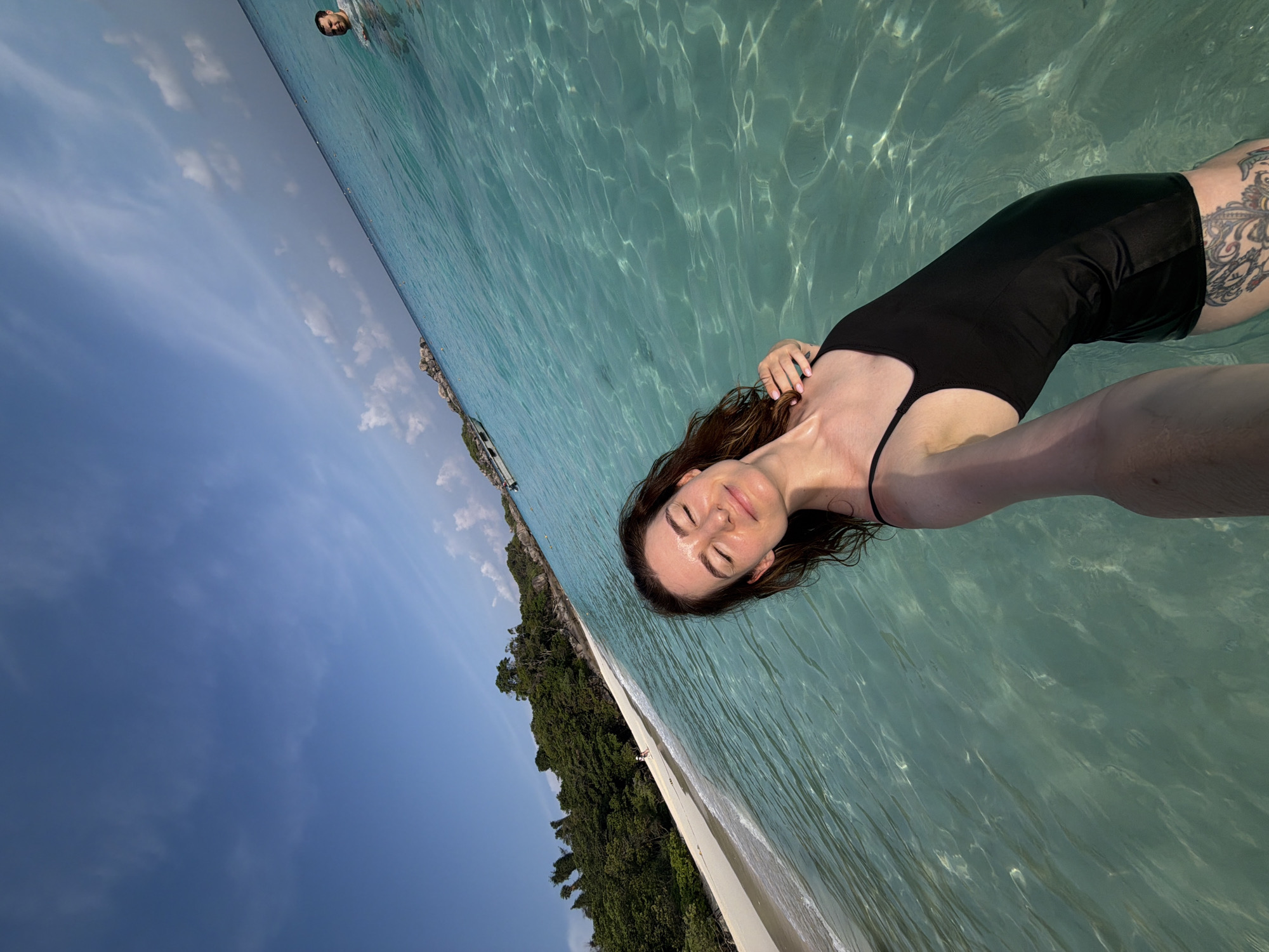 A female is depicted taking a selfie while standing in clear, shallow turquoise ocean water. She has closed eyes and a slight smile, with wet, dark brown hair. She wears a black one-piece swimsuit; a detailed tattoo is visible on her upper left thigh. Her left arm extends towards the camera; her right hand rests on her left shoulder. In the upper right background, a male is partially submerged in the water, facing left, with only his head and shoulders visible. To the left, a white sandy beach meets the water, backed by a lush green island covered with dense tropical trees. Distant dark rock formations rise from the water on the far right horizon, with a white boat visible further out. The sky is bright blue with scattered wispy white clouds. The scene is set in a coastal area of Lam Kaen, Thailand, under clear daylight conditions.