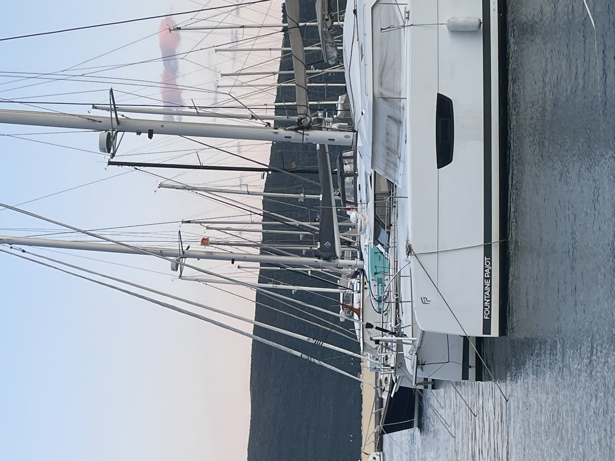 The image depicts a marina in Meljine, Montenegro, filled with numerous sailboats and catamarans docked side by side. The hulls of the boats are predominantly white. Many masts of the boats are visible, creating a forest of vertical lines against the backdrop of a hill, and what seems to be a plume of smoke. The catamaran closest to the camera has the name "FOUNTAINE PAJOT" printed on its hull. The water reflects the sky, creating a somewhat still surface alongside the boats.