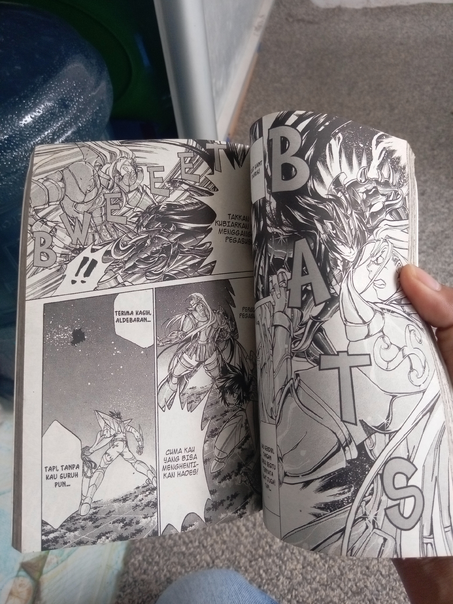 The image shows an open manga comic book, featuring a scene with several armored figures. On the left page, two characters are engaged in a fight, with large, bold letters "B" and "W" superimposed on the artwork. Text bubbles contain dialogue in Indonesian, including "TERIMA KASIH, ALDEBARAN..." and "TAPI TANPA KAU GURUH PUN...".
The right page displays a different panel with two characters in battle. Large, bold letters "B", "A", and "T" are visible across the page, partially obscuring the artwork. A character with long, dark hair and intense eyes is prominent, facing off against another armored figure. The text bubble on this page reads "PERGILAH PEGASUS!".
The background of the image suggests an indoor setting. To the left, a large blue water dispenser with condensation is visible, along with a green object that could be part of a container or furniture. The surface beneath the comic book appears to be a grey, textured carpet. A portion of a hand is visible on the right side of the image, holding the comic book open. The image was taken in Batam, Indonesia.
