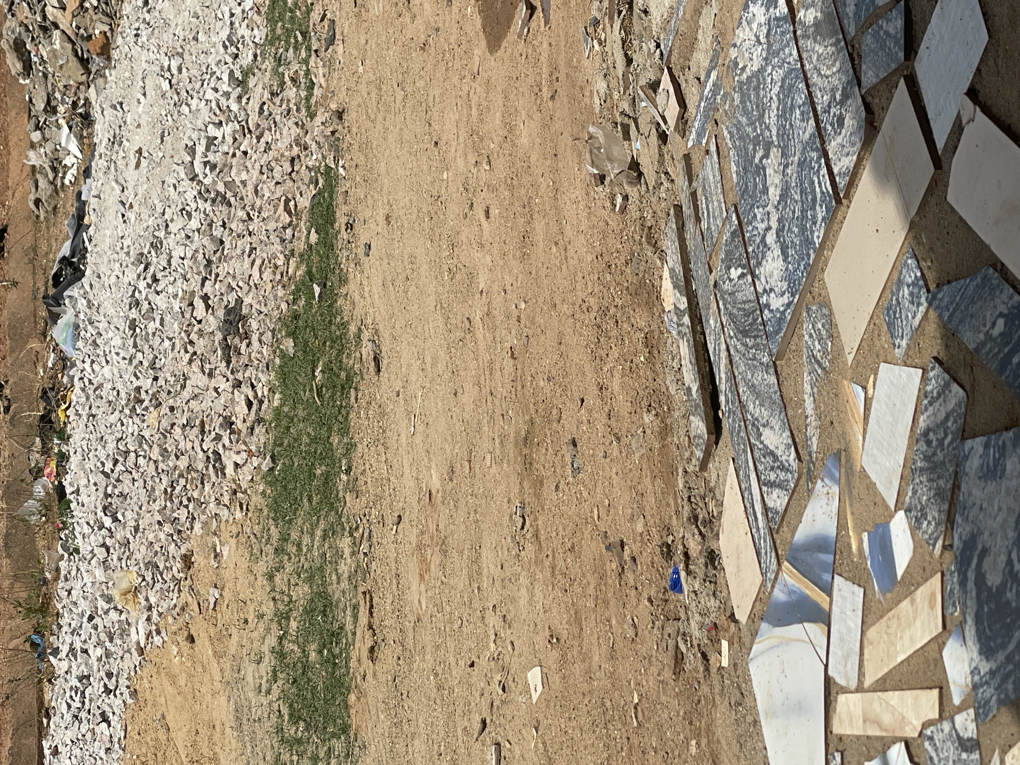 The image presents a ground-level, outdoor scene characterized by diverse textures and materials. The setting appears to be a dry, open area, possibly an undeveloped or neglected plot of land, consistent with its location in Narayi, Nigeria.
On the left side of the frame, there is a substantial pile of light-colored, jagged rocks or construction debris, interspersed with small pieces of trash, including plastic bags and dark plastic remnants. Adjacent to this rubble, a narrow strip of green grass grows.
The central part of the image is dominated by an expanse of bare, sandy-brown dirt, showing signs of being walked on or disturbed. This dirt area also contains scattered small pieces of debris, such as a crumpled clear plastic item and a blue plastic cap. More sparse patches of green grass are visible along the edges of this dirt path.
The right side of the scene features an informal mosaic created from irregularly shaped broken tiles and mirror pieces laid directly onto the dirt. The tiles display varying patterns, predominantly grey and white with dark, swirling veining, alongside some plain light beige or cream-colored pieces. Several mirror fragments are included, reflecting distorted views of the sky and surroundings.
No people or animals are visible, indicating a static scene. The bright, direct lighting suggests it is daytime, likely midday or early afternoon, with clear weather and no visible clouds. No discernible text is present in the image. The overall impression is one of an informal space with a mix of natural elements, construction waste, and repurposed materials.