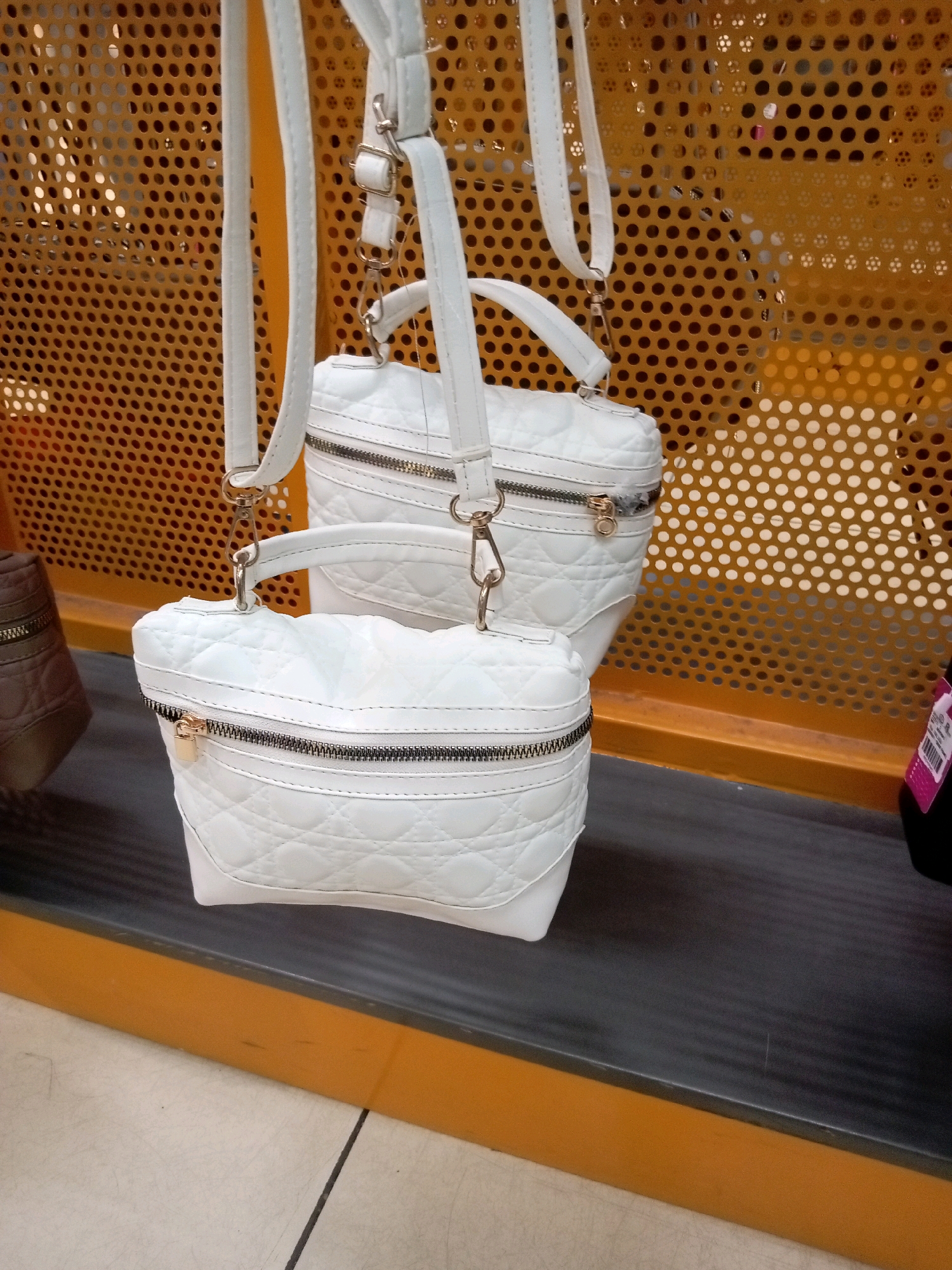 The image shows two white quilted handbags with gold-toned zippers and hardware, hanging from a perforated gold-toned display rack. The handbags appear to be made of faux leather and have a structured, boxy shape. The top handbag is slightly larger than the bottom one. A portion of a brown handbag is visible on the left side of the frame. The background is a blurred perforated gold metal sheet, likely part of a retail display. The foreground shows a dark grey shelf with an orange trim, and a tiled floor. The scene is indoors, possibly in a department store or boutique. There are no people visible in the image. Based on the context of the available information, this scene is likely located in Batam, Indonesia.