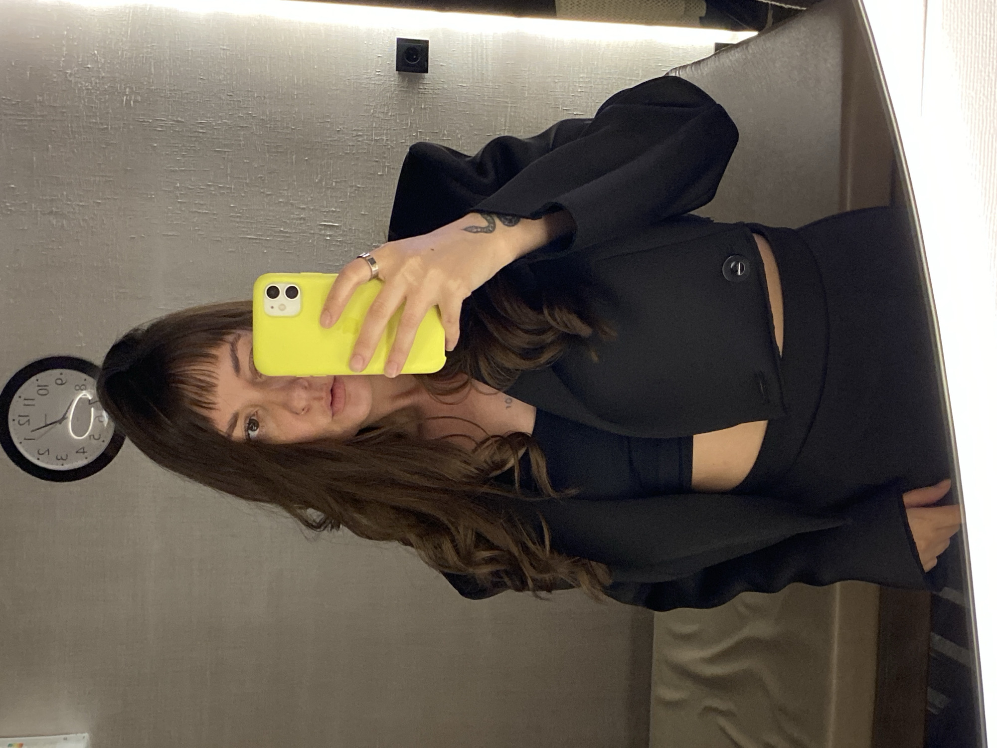 The image depicts an adult female engaged in a mirror selfie within an interior space, which the prompt identifies as being in the city of Votkinsk, Russia; however, no visual elements within the image confirm this geographical information.
The subject is a woman with long, dark brown, wavy hair and straight bangs, reflecting a neutral expression with slightly parted lips. She is wearing a black cropped blazer with prominent shoulder padding over a black crop top, revealing a sliver of her midriff. Her lower attire, also black, is visible as a skirt or trousers in the reflection. On her left wrist, she has a small, dark tattoo of a coiled snake, and a small numerical tattoo (possibly "10") is visible on her upper chest. She wears a silver-toned ring on the ring finger of her left hand.
She holds a bright yellow smartphone, identified by its dual rear cameras, obscuring the left side of her face. The background, a reflection in the mirror, features a light grey wall with a textured, possibly brushed or streaked, finish. An analog clock with a black frame and white face is visible in the upper left, displaying reversed numbers and indicating approximately 1:50 or 2:50 (actual time around 10:05 or 11:05). To the right, a vertical strip of light is reflected, and below it, a dark square object, possibly an electrical outlet or switch, is mounted on the wall. The lower portion of the image reflects a light-colored cushioned bench or seating area on the left and a darker, possibly upholstered surface on the right. The overall lighting is artificial and somewhat dim.
