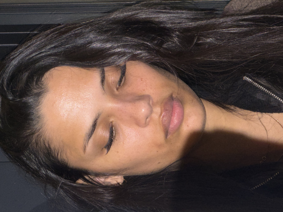 A young woman lies with closed eyes. Her dark, glossy hair frames her face, which is illuminated with a soft, warm light. She has smooth skin with a few freckles visible on her nose and cheeks. Her lips are full and slightly parted. A small gold earring is visible on her left ear. A thin gold necklace rests near her neck and what appears to be a black jacket is situated by the shoulder and neck.