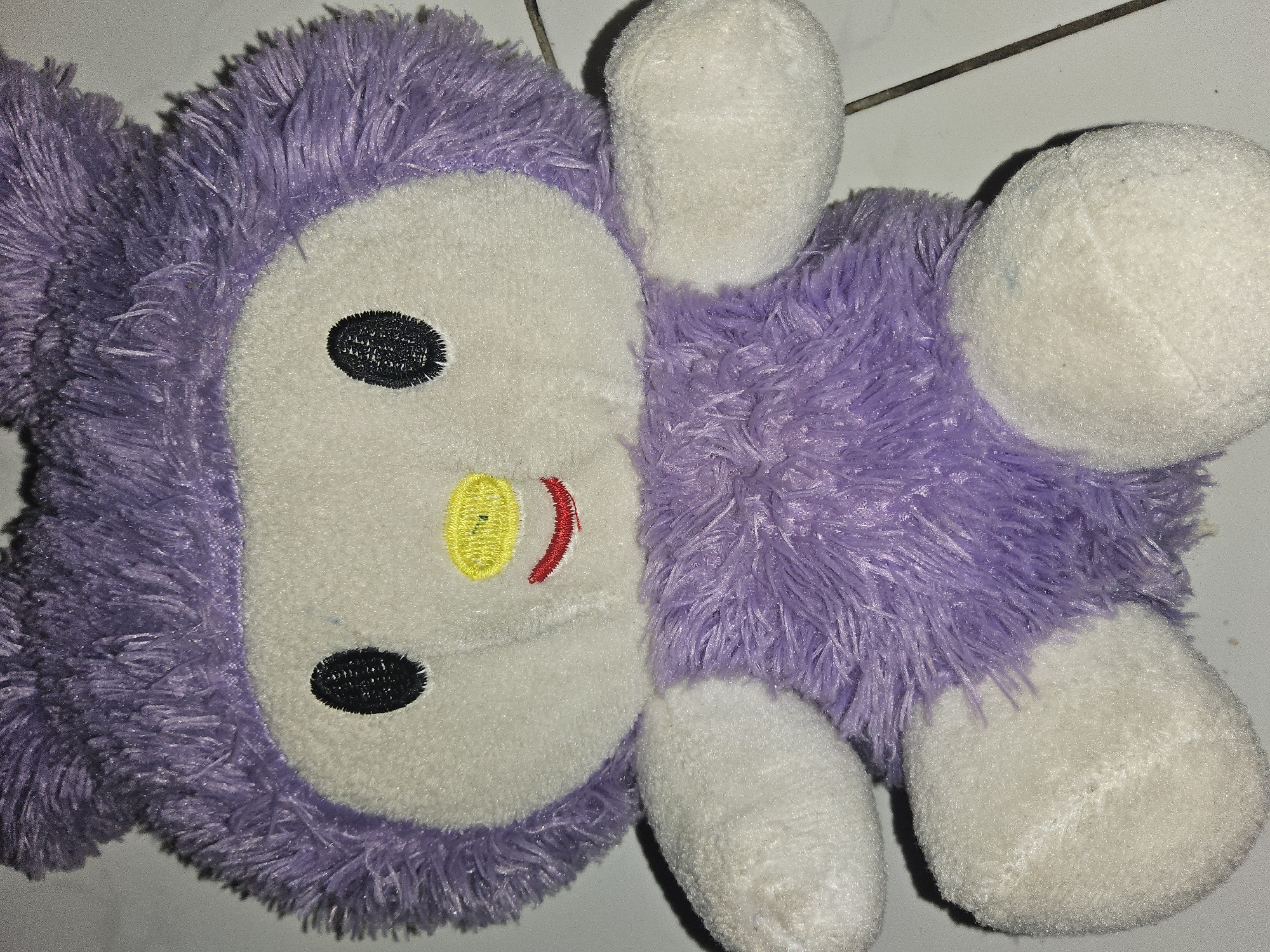 A purple and white plush toy is positioned in a sitting posture on a tiled surface. The toy features a white face with black embroidered eyes, a yellow embroidered nose, and a red embroidered mouth. Its body is covered in fluffy purple material, and it has white plush arms and legs. The background consists of a light-colored tiled floor, with a dark line running diagonally across the lower right portion of the image. There is a faint shadow cast by the toy on the floor. The scene is indoors. This image is not located in Payagaboh, Indonesia, but rather depicts a toy on a tiled surface.