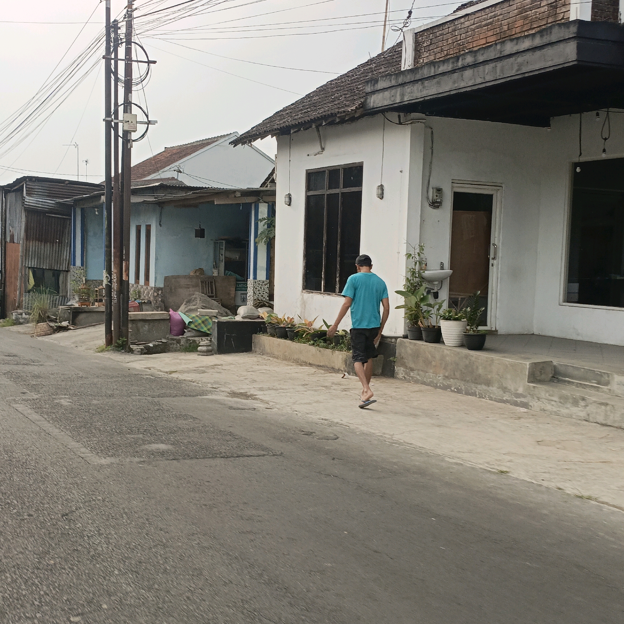 The image depicts a street scene in Salatiga, Indonesia, with a focus on a residential area. In the foreground, a gray asphalt road shows some wear and tear. A man wearing a blue t-shirt and black shorts, with a black cap, walks away from the camera on a concrete sidewalk. He is wearing flip-flops. The sidewalk runs alongside a white building with a dark awning and a tiled roof. This building has a large window with dark panes and an entrance door. Several potted plants are arranged in front of this building. To the left of the white building, there are other structures, including one with a corrugated metal roof and a light blue wall. A utility pole with numerous wires is situated between the road and the buildings. The overall environment appears to be overcast.