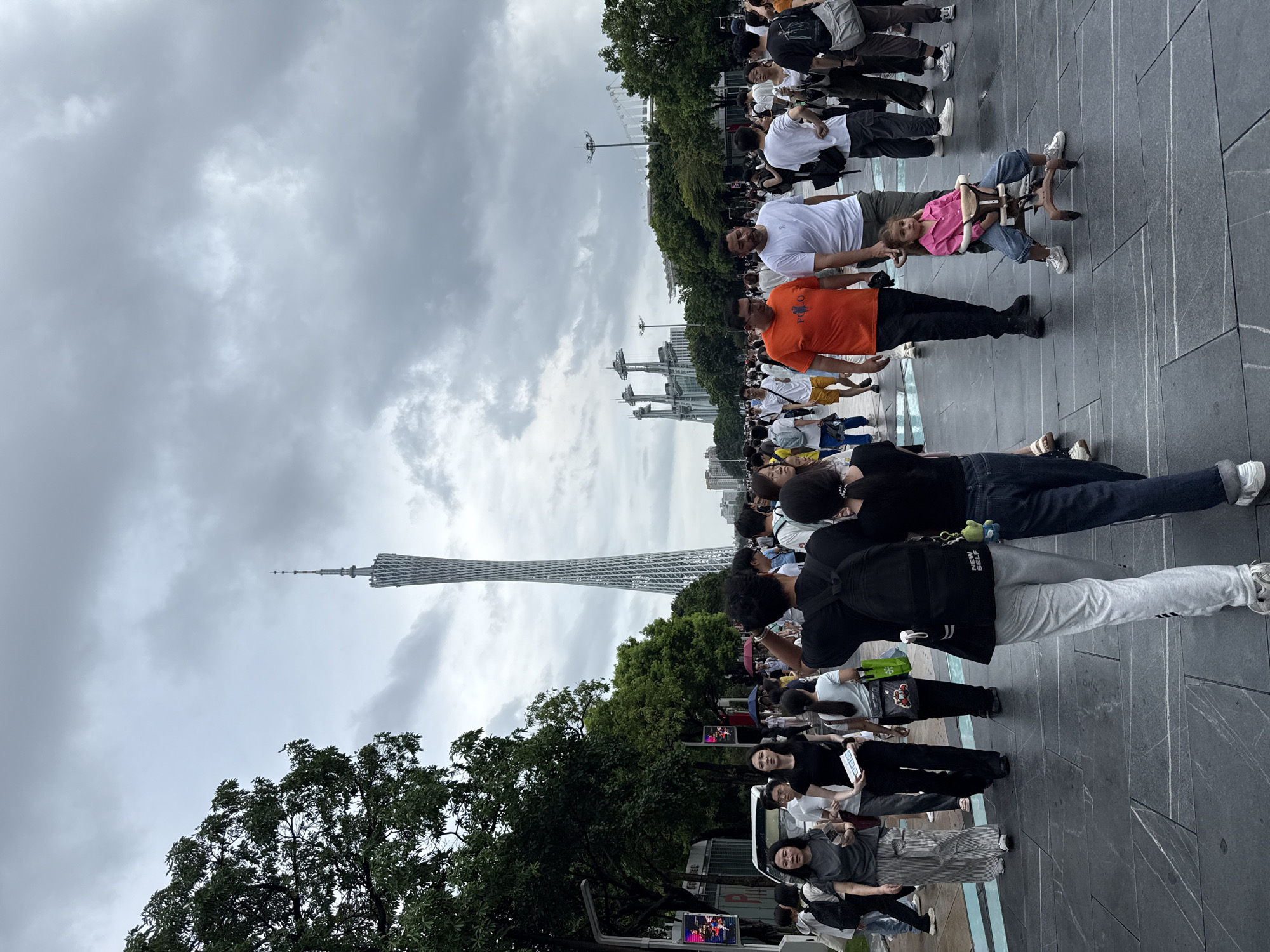 This image depicts a crowded outdoor scene in Tianhe District, China, with the Canton Tower visible in the background. A diverse group of people, including men, women, and a young child, are walking on a paved pathway. Some are carrying bags or wearing backpacks. The child is seated in a small, wheeled contraption. The sky is overcast with dense clouds, and trees line the left side of the pathway. Other structures, possibly part of a stadium or exhibition, are visible further in the distance. The overall atmosphere suggests a public gathering or busy pedestrian area.