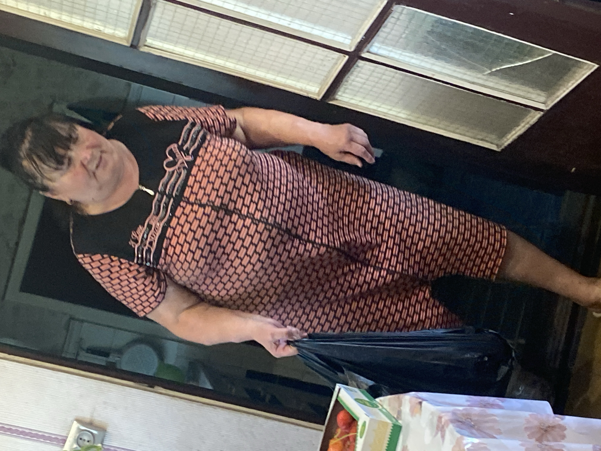 An elderly woman with dark hair and bangs, wearing a short-sleeved pink and black patterned dress, stands in a doorway. She is holding a black plastic bag in her right hand. To her left, a table covered with a floral tablecloth holds a white box with green trim, containing red fruit, possibly cherries. The background shows a kitchen with a white wall outlet, a pan hanging on the wall, and a dark doorway. The scene appears to be located in Vvedenka, Ukraine.