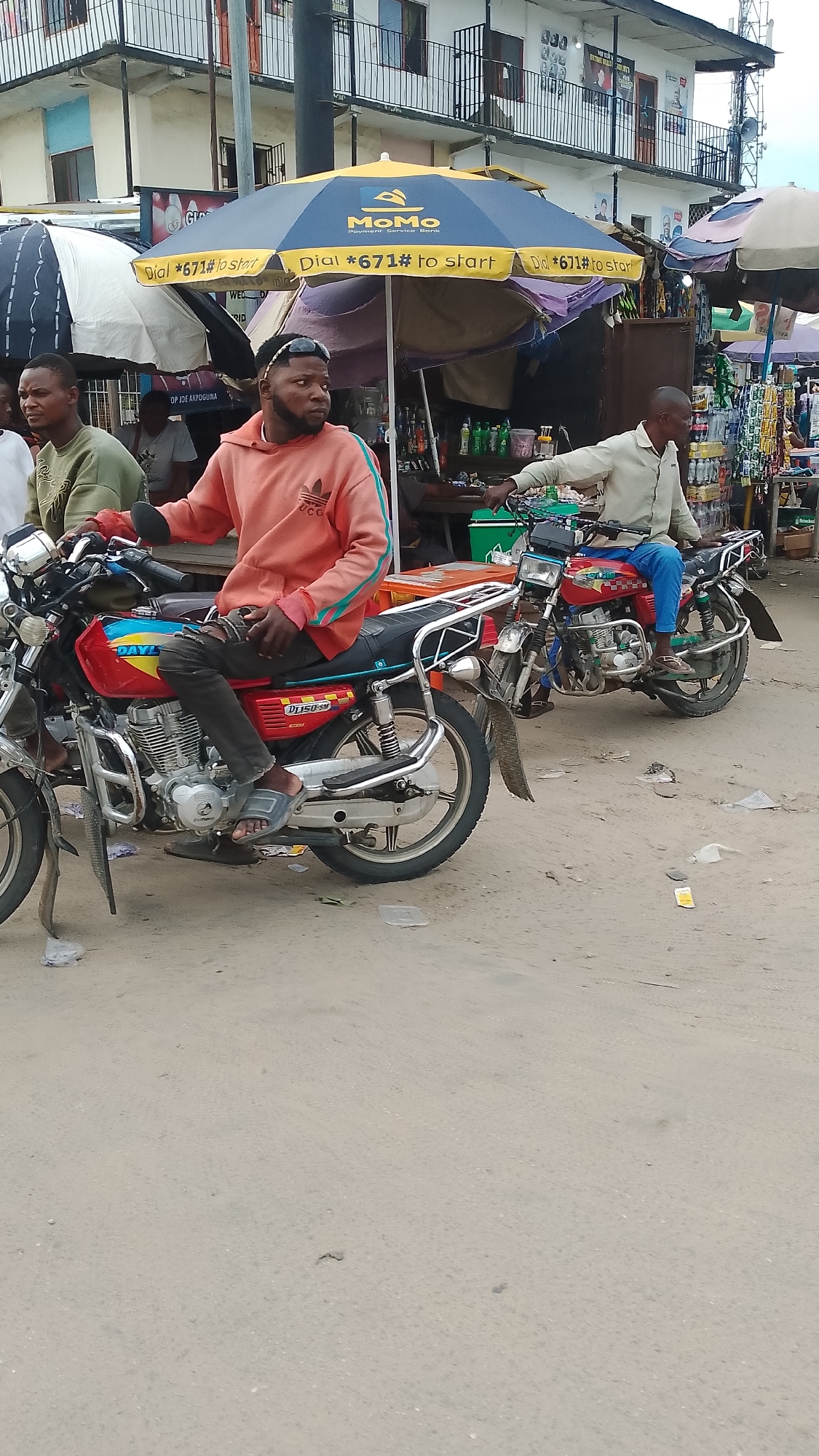 The image depicts a street scene in Agbarho, Nigeria, with two men on motorcycles. The man on the left, wearing a green sweater, is partially visible. The man in the foreground, seated on a red motorcycle, is wearing an orange hoodie and dark pants. He has a beard and is wearing sunglasses on his head. A blue and yellow umbrella with the "MoMo Payment Service Bank" logo is open above a stall in the background. The stall appears to be selling various items, including drinks. Another man in a light-colored shirt and blue pants is on a red motorcycle to the right. The ground is dusty and appears to be a dirt road or a poorly paved street. Buildings are visible in the background, with balconies and windows. The overall impression is a bustling marketplace environment.