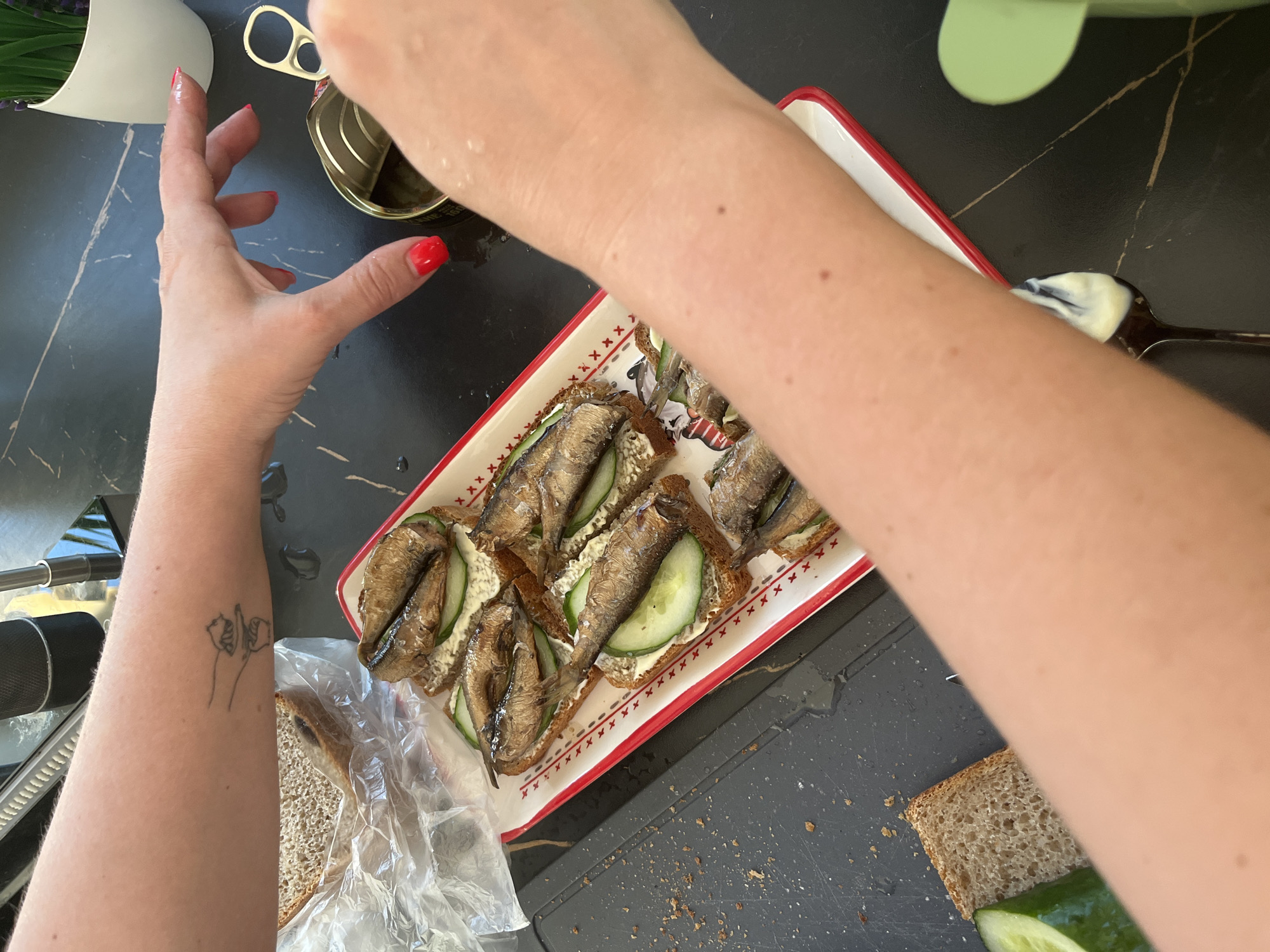 A top-down shot shows a person preparing sandwiches, likely in Новая Чернушка, Russia. The ingredients are arranged on a black countertop with light-colored marble-like veining. A white rectangular plate with a red border holds several open-faced sandwiches, each consisting of a slice of brown bread topped with a white spread, cucumber slices, and smoked sprats. A can of sprats sits open next to the sandwiches. The person's hands are visible, with one hand opening the can of sprats and the other reaching for the sandwich plate. Visible on the forearm is a tattoo of two stick figures. A plastic bag containing sliced bread sits near the plate. A spoon with a white substance on it is also visible.