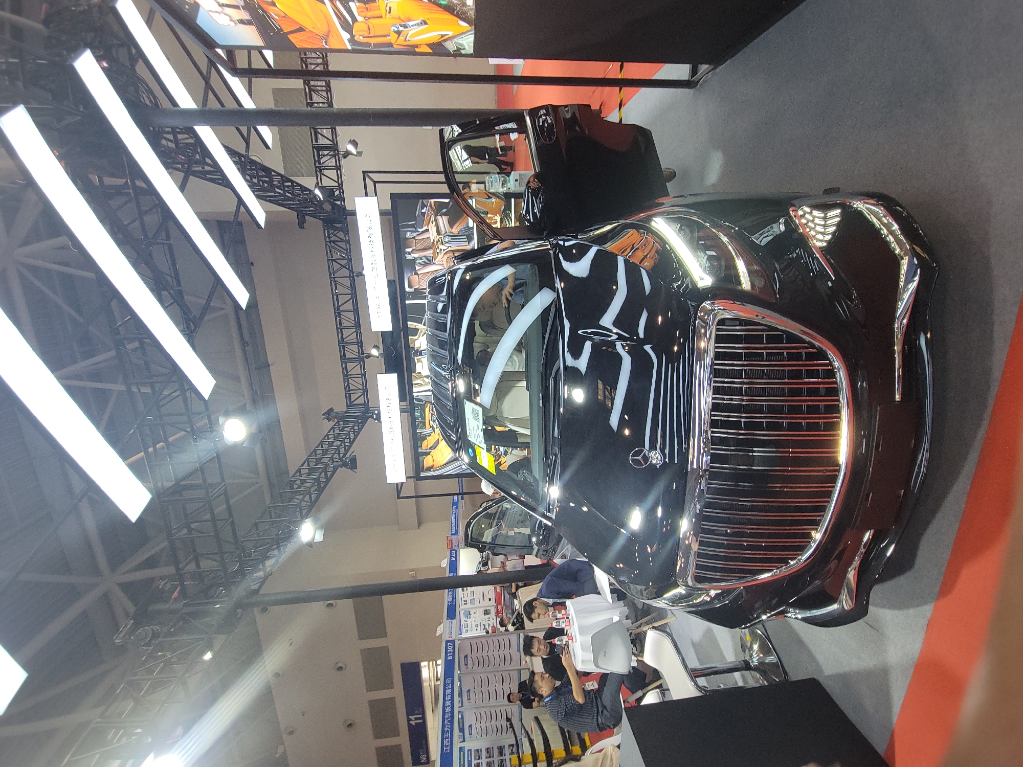 The image shows a black luxury car with chrome grill on display at an event. Several people are seated around the car, seemingly engaged in a meeting. A display board with various products is visible in the background. The location is the city of Liangjiang, China, based on the Chinese writing on a sign near the car. Overhead lighting and support structures are also visible.
