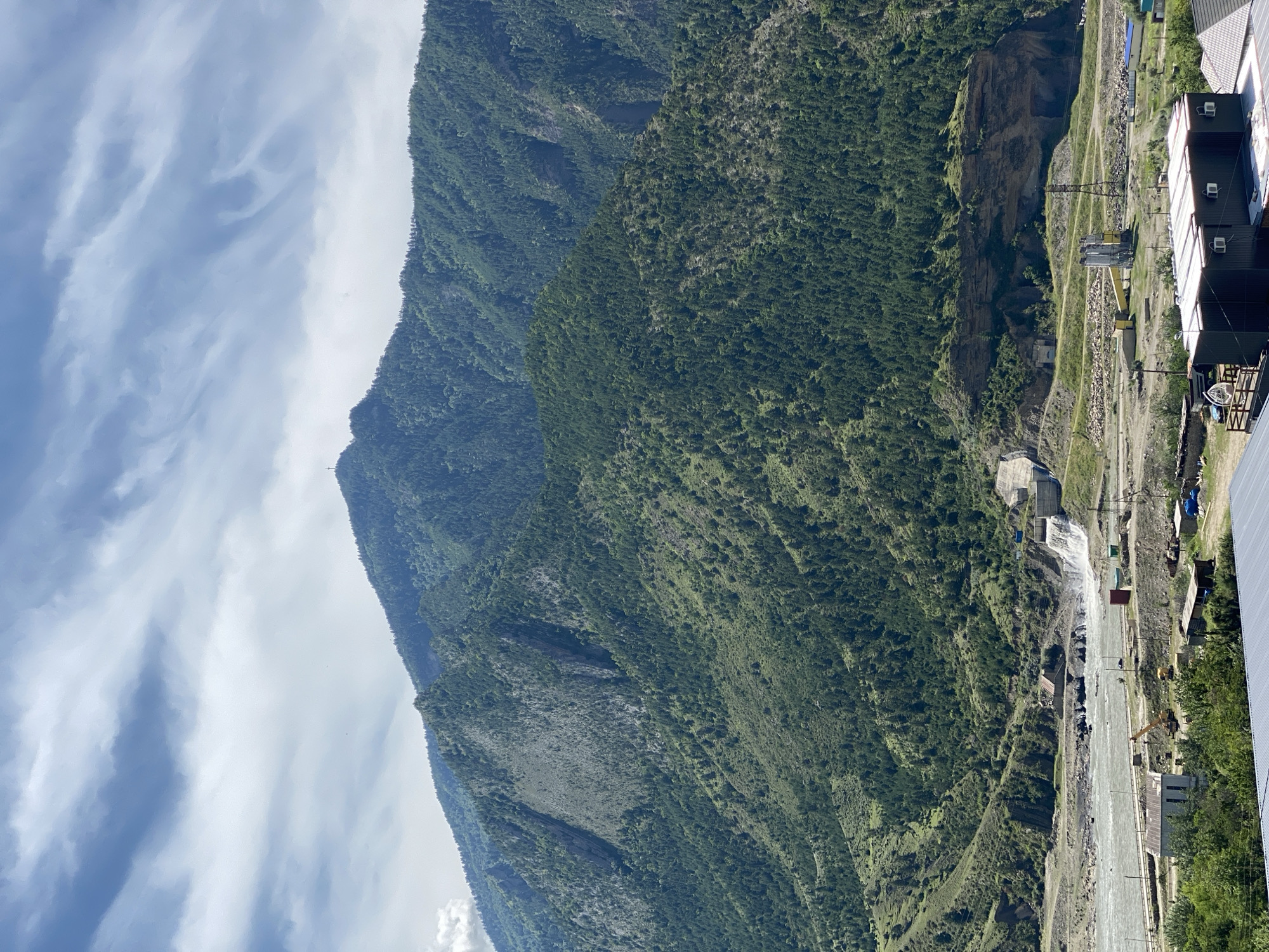The image depicts a mountainous landscape with a river and a dam in the foreground. The mountains are covered in dense green vegetation, indicative of a forested area. In the mid-ground, a river flows from left to right, with a dam on the right side of the river generating a waterfall. The dam appears to be a concrete structure. Near the dam, there are some industrial buildings and construction equipment, including a yellow crane. The foreground shows a few smaller buildings and some trees. The sky is cloudy, with streaks of light blue visible. The image appears to be taken from an elevated position, offering a panoramic view of the scene. While the exact location in Chalda, Russia, is not explicitly identifiable without further context, the mountainous and riverine environment suggests a region with such geographical features. No people are visible in the image.