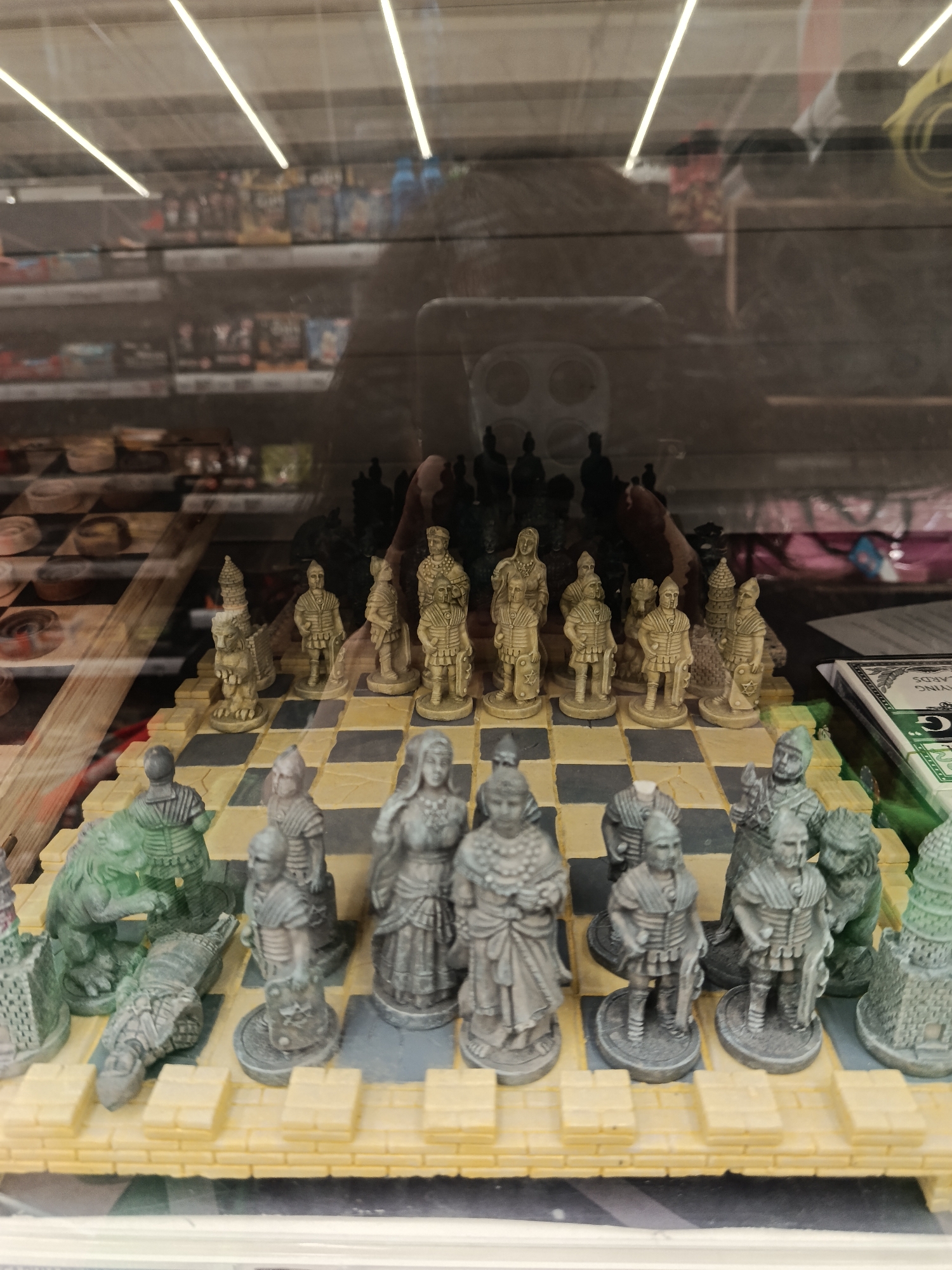 The image shows a chess set with Roman-themed pieces displayed inside a glass cabinet. The chess pieces are made of stone material and are arranged on a checkered board. The pieces are divided into light and dark colors, indicating opposing sides. The environment appears to be a retail setting, given the shelves filled with various items in the background behind the reflection on the glass. I am unable to determine any of the items on the shelves behind the chess set. I am unable to determine the specific location in Moscow, Russia, based on the provided image.