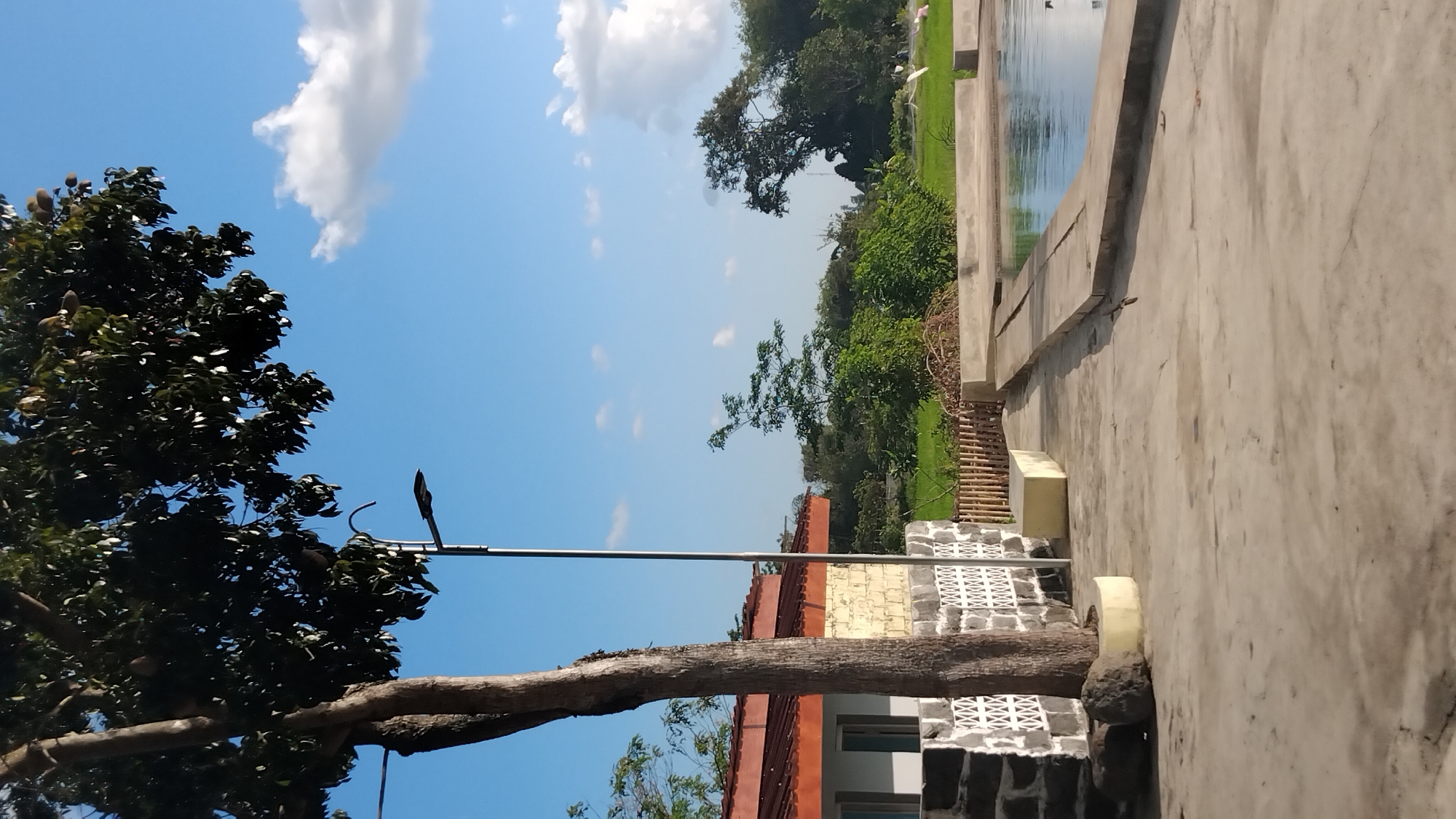 The image depicts an outdoor scene in Ngaglik, Indonesia. In the foreground, a paved area with some cracks leads to a swimming pool filled with blue water. The pool has a concrete border. To the left, a building with a reddish-brown roof and a light-colored brick wall is visible, adjacent to a stone wall with decorative patterns. A tree with dark green foliage stands prominently next to the building. Behind the building and pool, lush green vegetation and trees form the background. The sky is blue with scattered white clouds. A street lamp is positioned near the tree and building. The overall scene suggests a tranquil, possibly residential or recreational, environment in a tropical location.