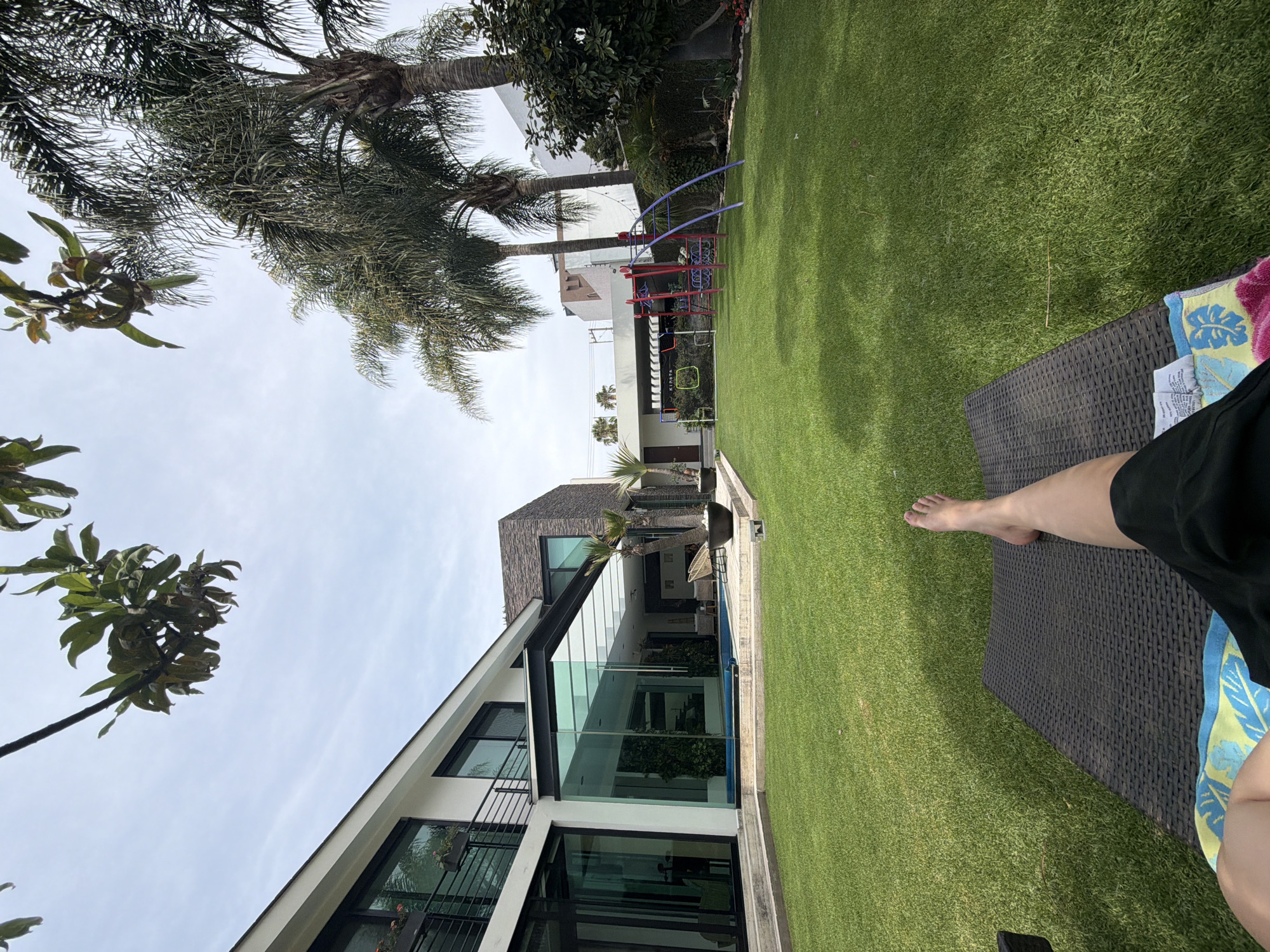 The image shows a person reclining on a dark brown wicker chaise lounge, partially visible from the legs down. The scene takes place in a lawn area with artificial green turf. A modern house with large glass windows is visible in the background. A small pool is located near the house. The sky is overcast. Palm trees are present. A play structure is located further away in the yard. The location is not identified beyond a city and state labeled as "Unknown, Unknown".