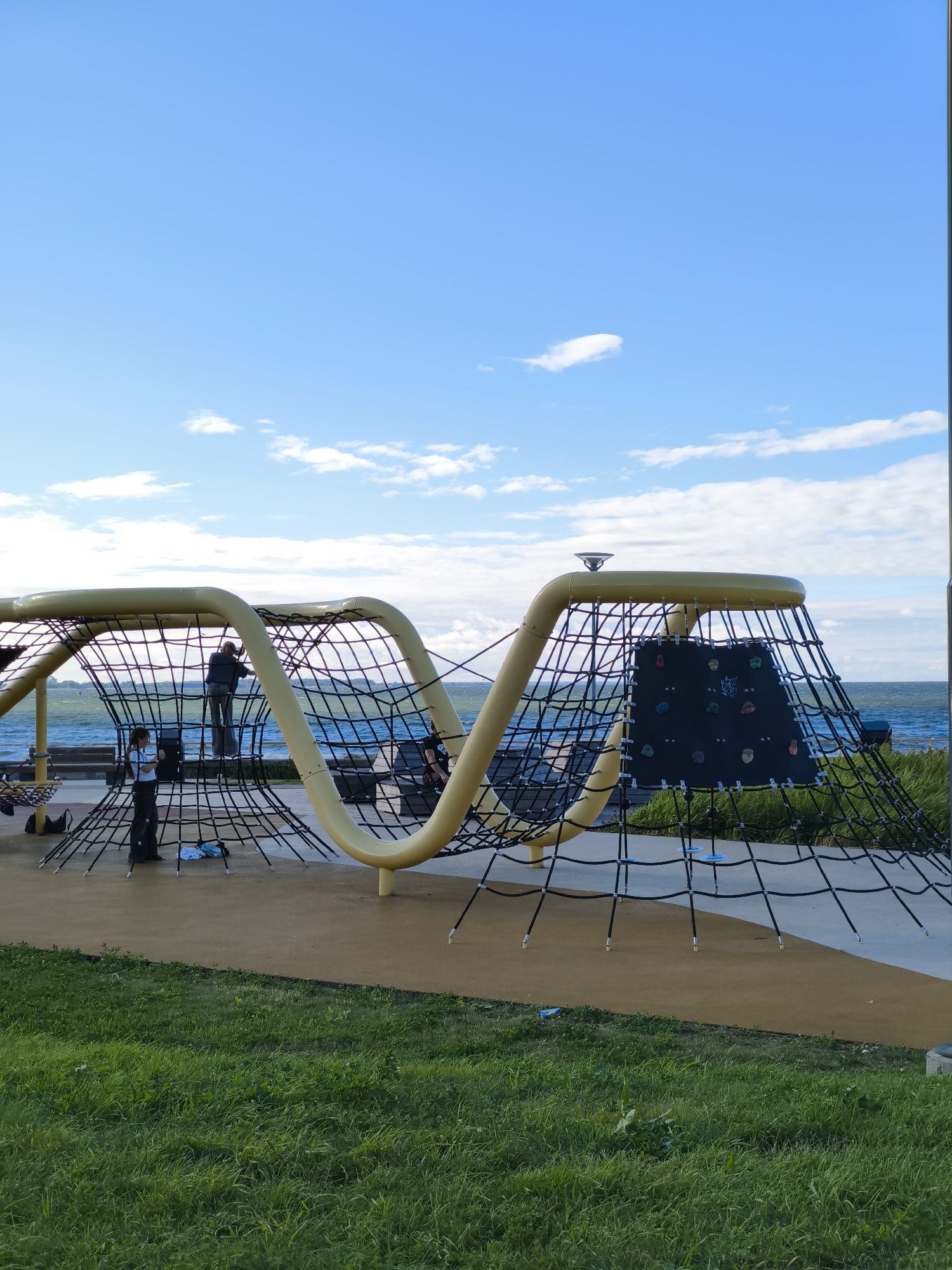 The image depicts a modern outdoor playground with a prominent yellow, curved structure and a black rope net climbing area. A few individuals are present on the playground. One person, viewed from the back, is standing on the rope net structure, facing the water. Another person, a female, stands on the ground to the left of the structure, looking upwards. A third person is partially visible on a bench to the right.
The playground is situated near a large body of water, likely a bay or sea, with waves visible. The water is blue and stretches towards the horizon under a bright blue sky dotted with scattered white clouds. Beyond the water, a distant shoreline with buildings can be seen.
The foreground consists of green grass and a light brown, rubberized surface which forms the playground's safety surfacing. The playground equipment itself features a climbing wall with colorful holds attached to a black panel on the right side of the structure. The yellow structure appears to be made of metal tubing, forming a kind of elevated walkway or climbing frame.
This scene is consistent with a public recreational area, possibly in a coastal part of Tallinn, Estonia, given the water and the visible hints of urban landscape in the distance.