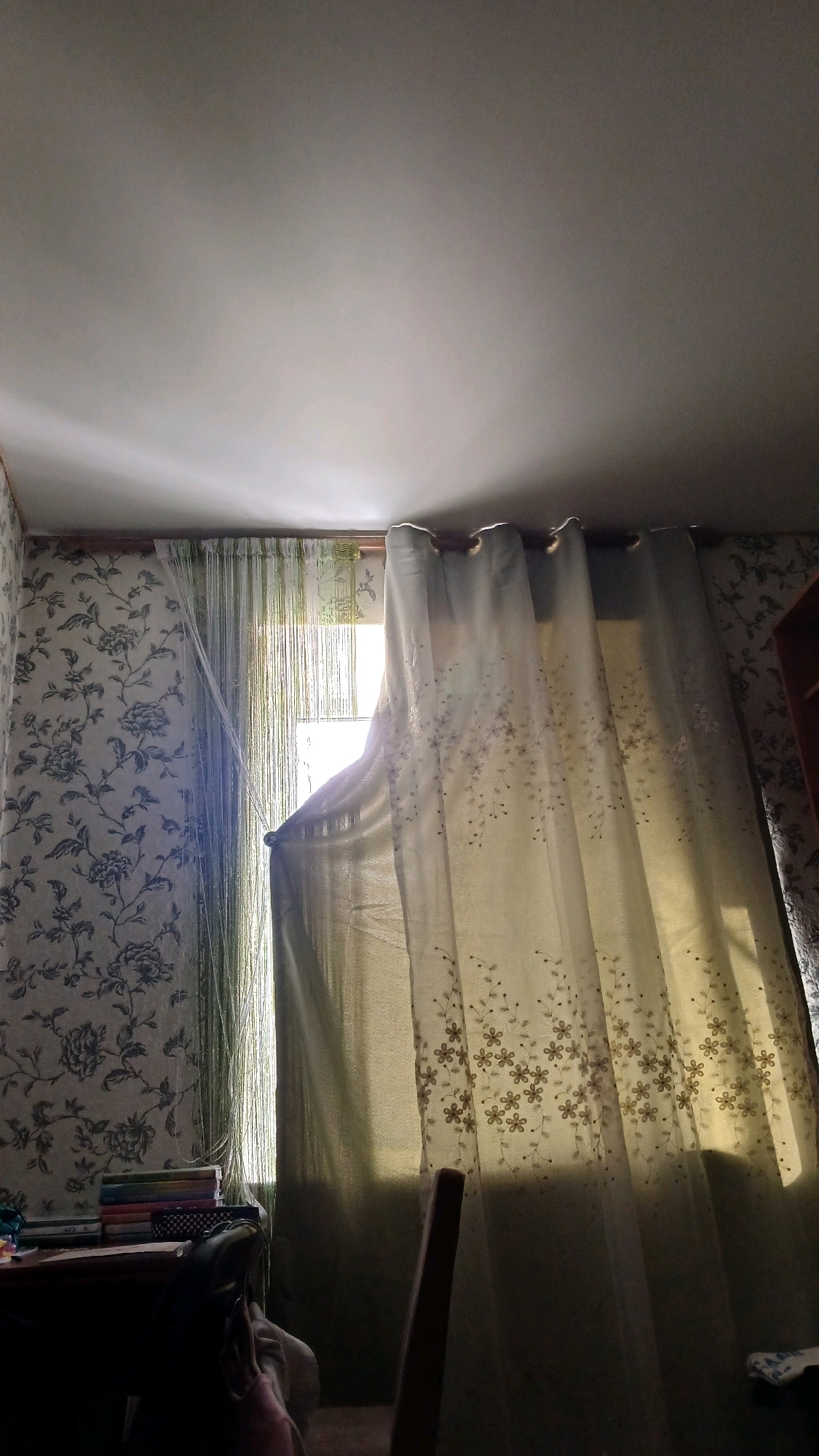 The image depicts an interior scene with a window covered by curtains. Sunlight streams through the window, illuminating the room and creating shadows. On the left, floral wallpaper covers the wall. To the right, a sheer curtain with a floral pattern is partially drawn, revealing a beaded curtain behind it. In the lower left corner, a desk with a stack of books is visible. A chair back is partially visible in the foreground. The ceiling is light-colored and shows some light reflections. The scene appears to be a domestic interior, possibly a living room or bedroom. There is no visible indication of the specific location being Haivoron, Ukraine.
