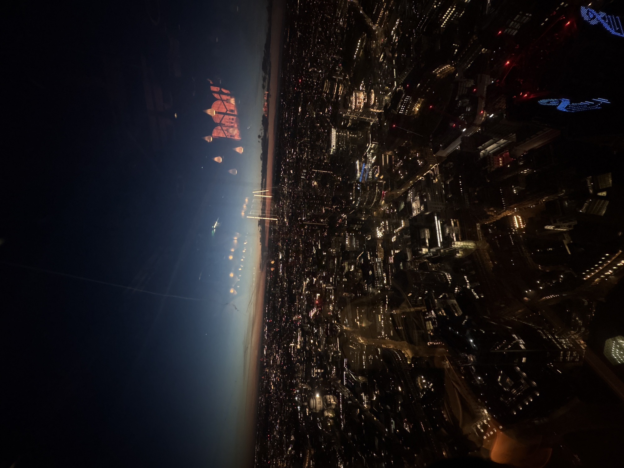 An aerial view of the city of London at night. The city is illuminated with a dense network of lights from buildings, streets, and landmarks. The skyline is a mix of low-rise and high-rise structures. The River Thames can be seen snaking through the city, reflecting the city's lights. In the sky above, there is a reflection of the interior of the airplane, including lights, an emergency exit sign, and window shades. The Shard and other prominent buildings are visible landmarks within the cityscape.