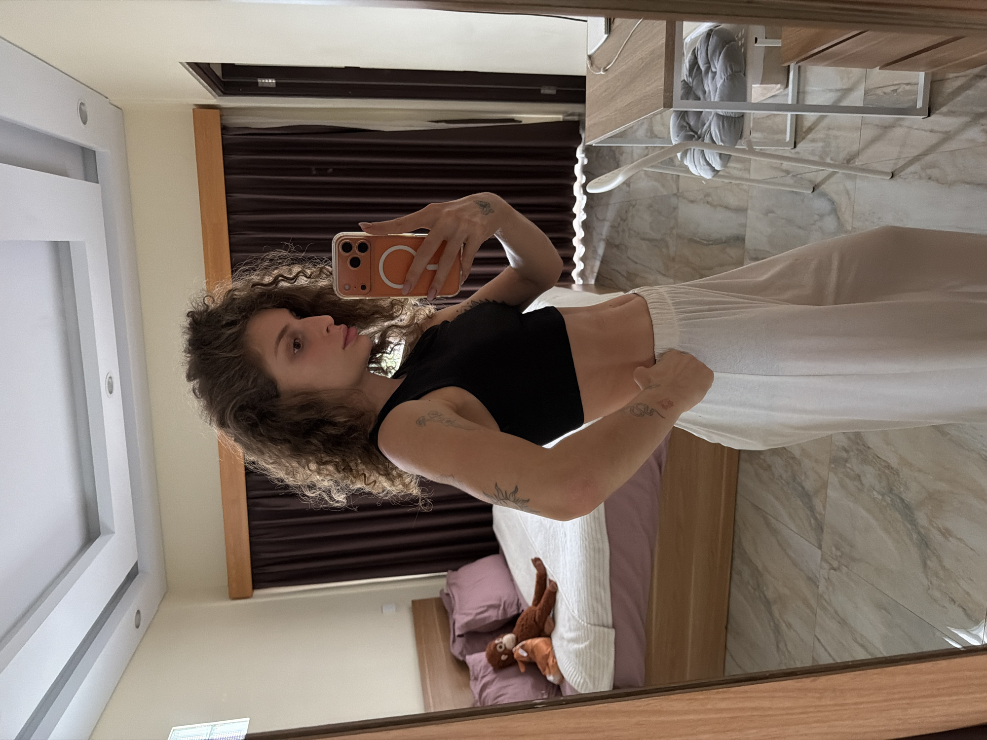 A young woman with curly brown hair stands in a bedroom in North Kuta, Indonesia, taking a selfie in a mirror. She is wearing a black crop top and loose white pants. The room has a bed with purple bedding and two stuffed animals, a desk with a chair, and a door. The floor is tiled. The overall impression is a casual, domestic setting.