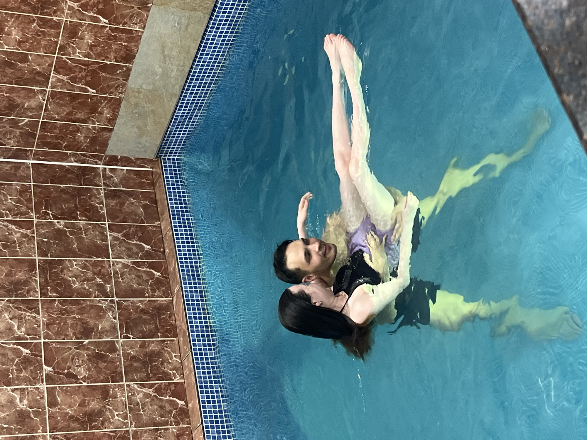 An overhead, slightly angled shot captures two individuals, a man and a woman, embraced and floating in a tranquil indoor swimming pool. The man, positioned with his back mostly to the viewer, has short dark hair and a gentle smile as he looks upwards and slightly right. He wears a dark top. The woman, with long dark hair, rests her head against his shoulder, looking towards him with a soft expression. She wears a dark, possibly sequined, halterneck top.
Their bodies are partially submerged in the clear, blue water. The woman's legs are gracefully extended upwards and slightly out of the water, while the outlines of the man's legs are visible beneath the surface. The pool's edge is lined with small blue mosaic tiles. Surrounding the pool, the floor features a border of light-colored rectangular tiles, adjacent to a larger area paved with dark brown, marble-patterned tiles with white grout lines. The setting suggests an indoor facility, possibly in Chaikovsky, Russia, with artificial lighting illuminating the scene. Both individuals display expressions of contentment or enjoyment. There is no visible text in the image.