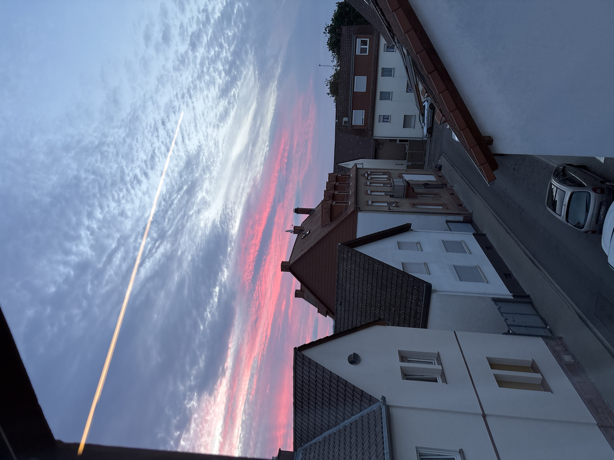 The image depicts a residential street in Viernheim, Germany, during a sunset. The sky is a gradient of blue and pink, with a thin, bright orange contrail stretching across it. Several houses with red-tiled roofs and white facades line the street. On the street, two cars are parked: a silver SUV and a silver hatchback. The foreground is dominated by the roof and upper facade of a white house. The overall scene is one of a tranquil evening in a European town.
