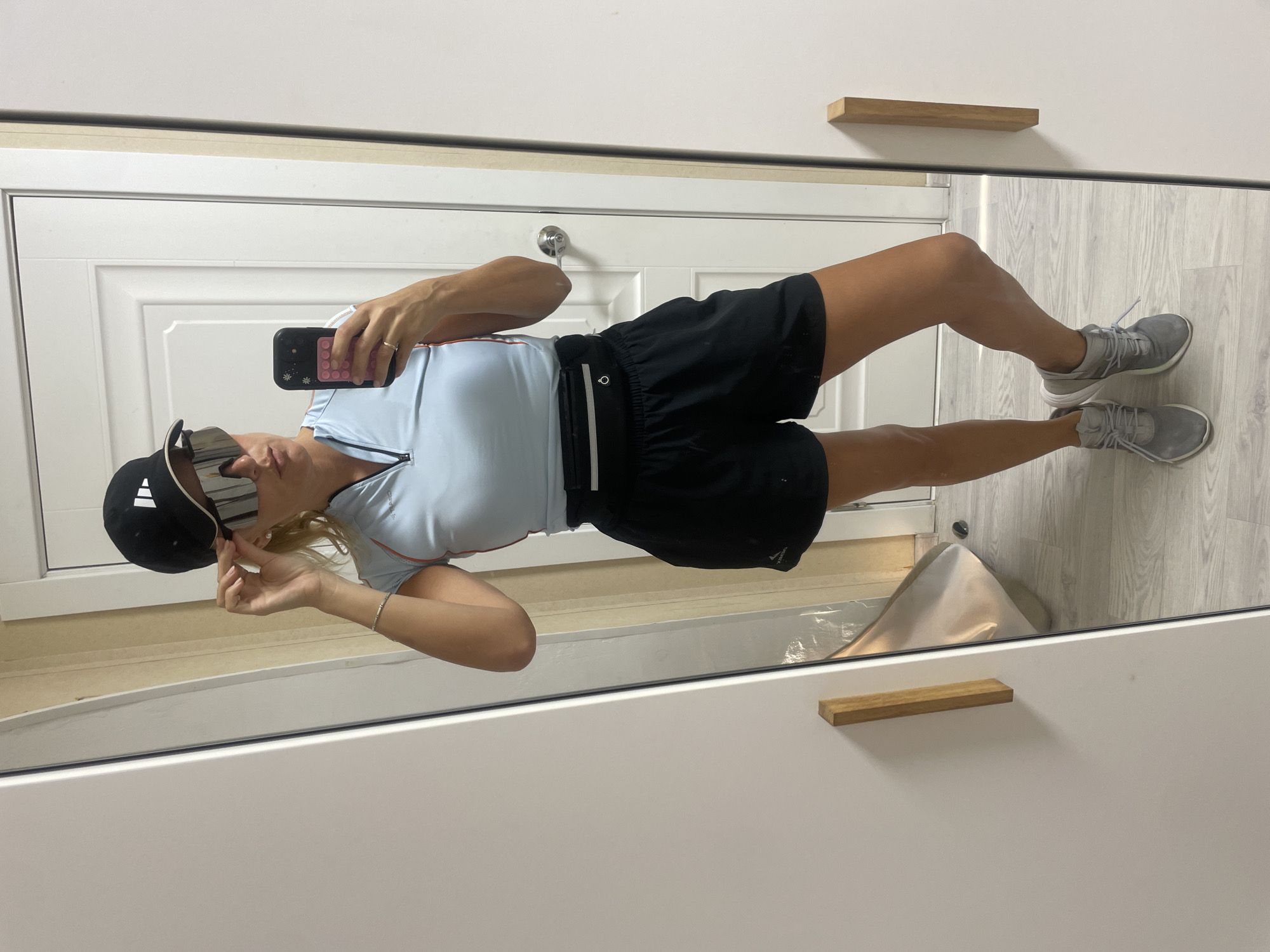 A person, likely female, stands indoors taking a mirror selfie. She is dressed in athletic attire, featuring a light blue short-sleeved top with a quarter-zip front and orange/pink piping, paired with black athletic shorts that have a subtle "XTERRA" logo. A black fanny pack or belt bag is secured around her waist. She wears a black baseball cap with a white Adidas logo, large reflective silver sunglasses, and grey athletic sneakers.
Her left hand holds a black smartphone with a distinctive pink pop-it style case, and a ring is visible on her finger. A thin bracelet adorns her right wrist. The image is a reflection captured in a large mirror. The background seen in the reflection consists of a white paneled door with a silver doorknob. The floor is covered in a light-colored wood or laminate material. In the immediate foreground of the reflection, a white cabinet or dresser with rectangular wooden handles is visible. A white cloth or bag lies on the floor in the reflected space. The setting appears to be an indoor room in Baranovka, Russia, with no specific time of day discernible.