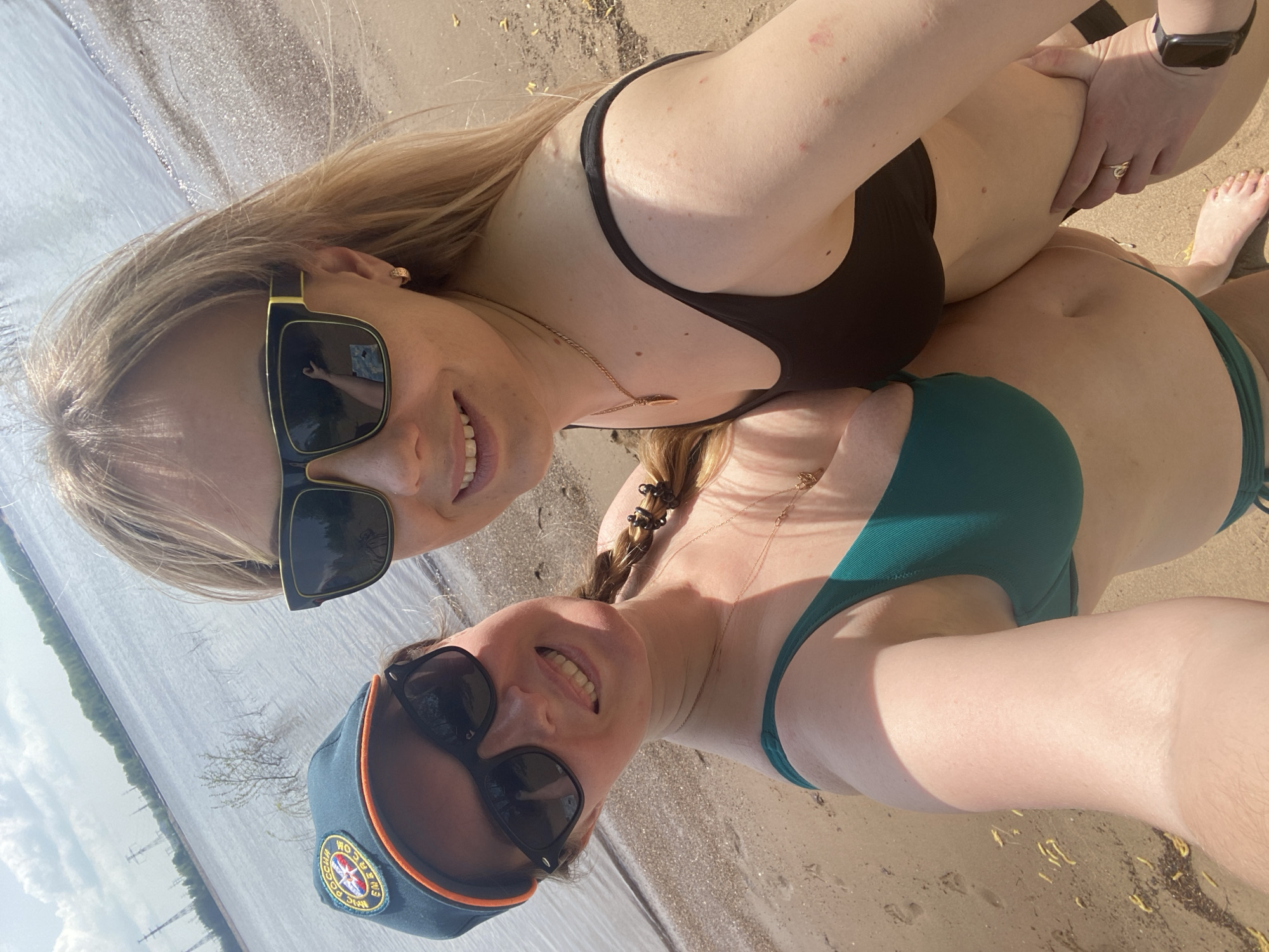 Two women are posing for a selfie on a beach. The woman on the top wears a black bikini and sunglasses, while the woman on the bottom wears a green bikini and a hat. The background shows a body of water and some vegetation. It is not possible to confirm if the image was taken in the city of Chaikovsky, Russia based on visual evidence alone.