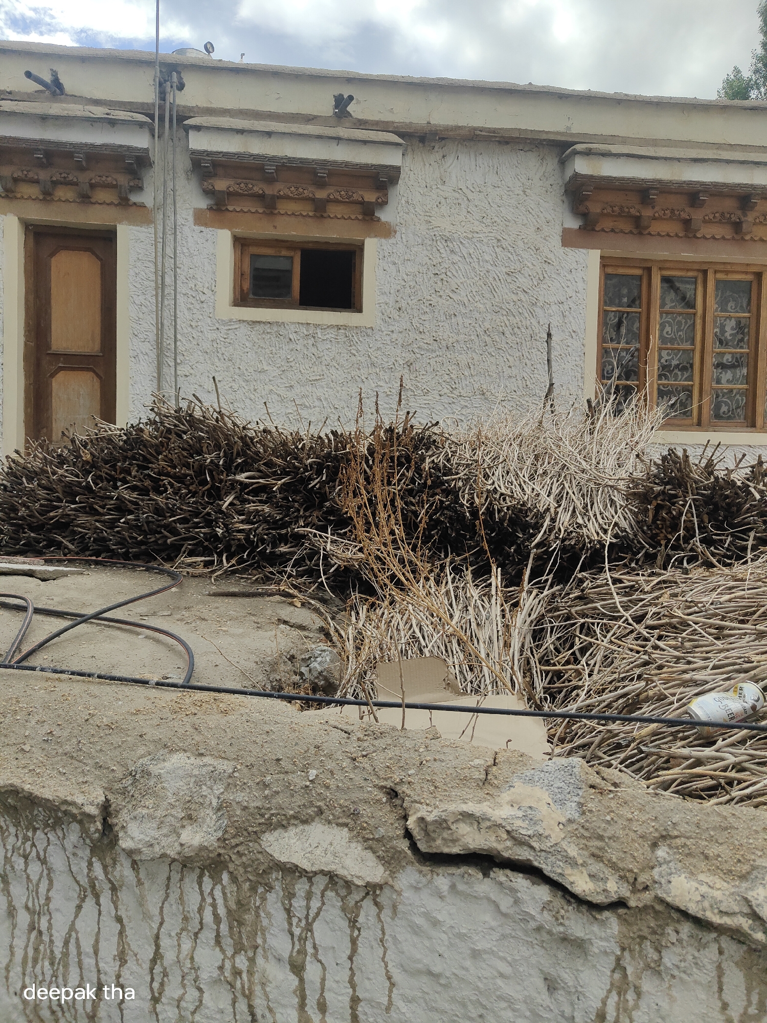The image shows a white, textured building with two windows and a door. The windows have ornate wooden frames and the glass panes have decorative patterns. A pile of dry branches and twigs is stacked in front of the building. A black hose is coiled on the ground in front of the pile of branches. In the foreground, there is a cracked concrete wall with water stains running down its side. The sky is overcast. The name "deepak tha" is visible in the lower left corner of the image. This scene is likely located in the city of Leh, India, given the architectural style and the mountainous terrain implied by the dry vegetation.