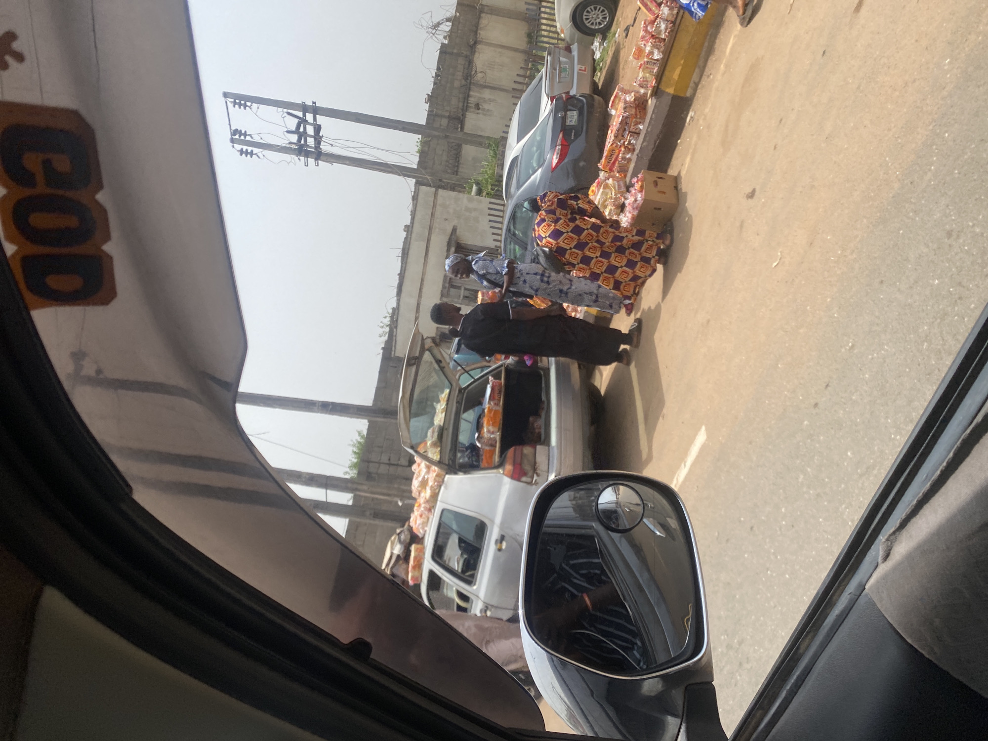 Captured from inside a vehicle, the image shows a street scene in Alakia, Nigeria. A white commercial vehicle with its trunk open is parked alongside a road filled with various goods. Several people, including at least two women in patterned clothing, are interacting with the merchandise. A grey car is also parked near the vendor's space. The environment appears to be a bustling street with buildings and utility poles visible in the background. The overall setting suggests a typical roadside market scene in Alakia.