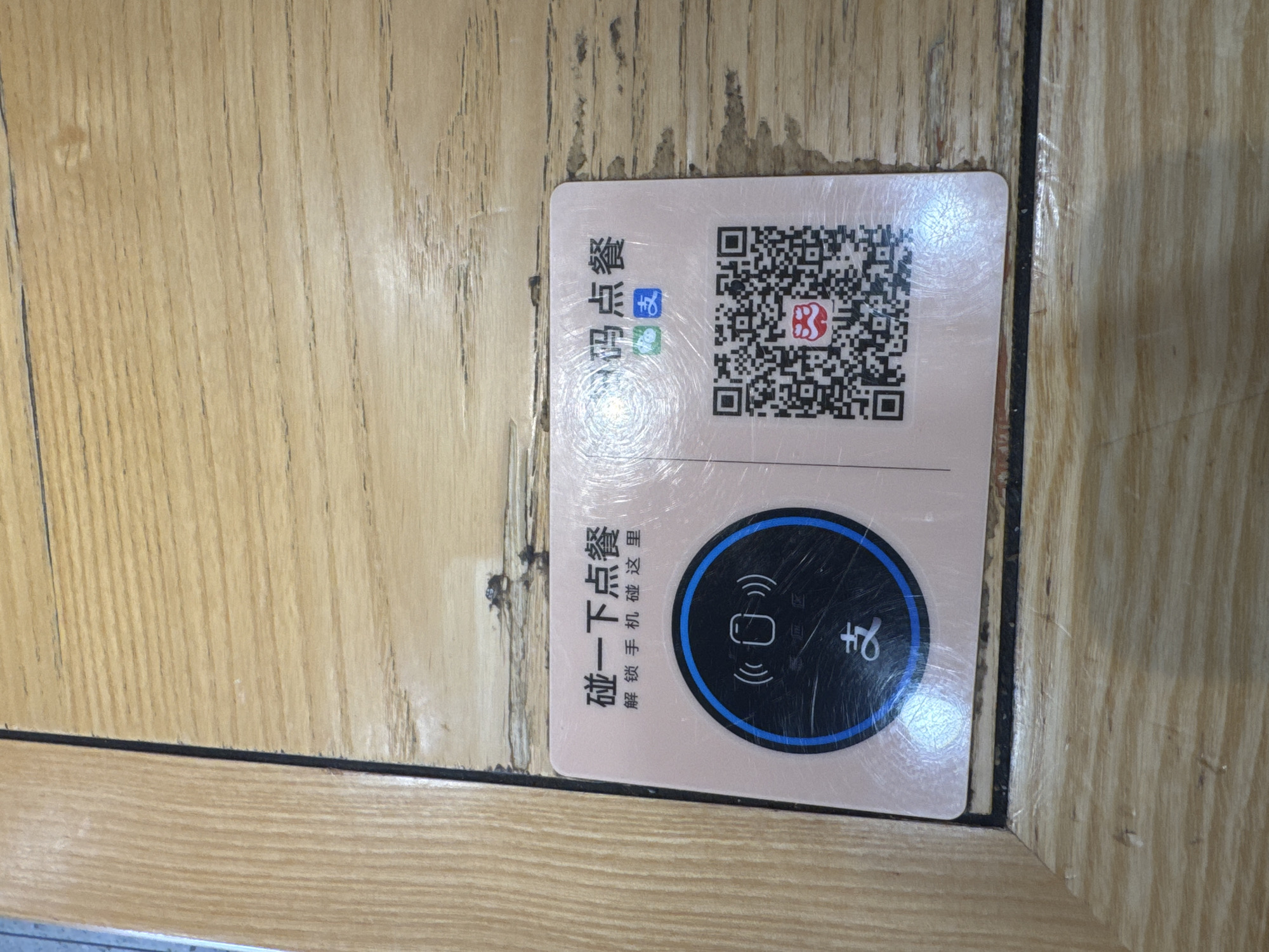 A light pink card with Chinese text and two QR codes is placed on a wooden table. The card has text that reads "碰一下点餐" (touch to order food) and "解锁手机碰这里" (unlock phone touch here). Below this text, there is a circular area with a stylized icon of a phone and radiating waves, suggesting NFC functionality. Below this circle, the Chinese character "支" is visible. On the right side of the card, there is a QR code. Below the QR code, there is a smaller red and white logo. The wooden table has a distinct grain pattern. The image appears to be taken in Dadong District, China.